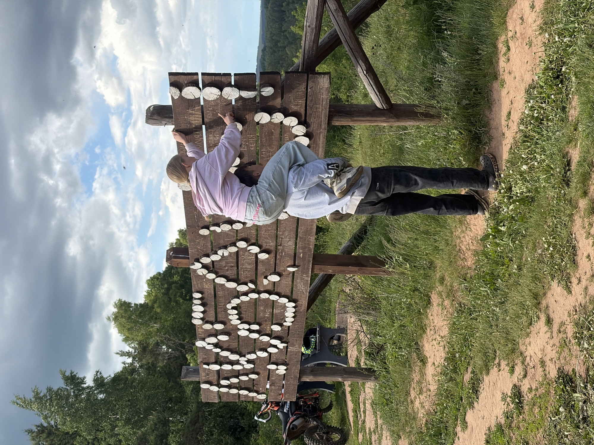 The image shows a wooden sign with the words "LESO GRAD" spelled out in white wood slices, located in what appears to be a rural area. Two individuals are on the sign, one lying on their back and the other lying atop them, reaching towards the word. A motorcycle is visible at the bottom of the sign, indicating this area is a park or outdoor recreation site. The background consists of greenery, indicating a forest. Based on the image request, the location is Костоваты, Russia.