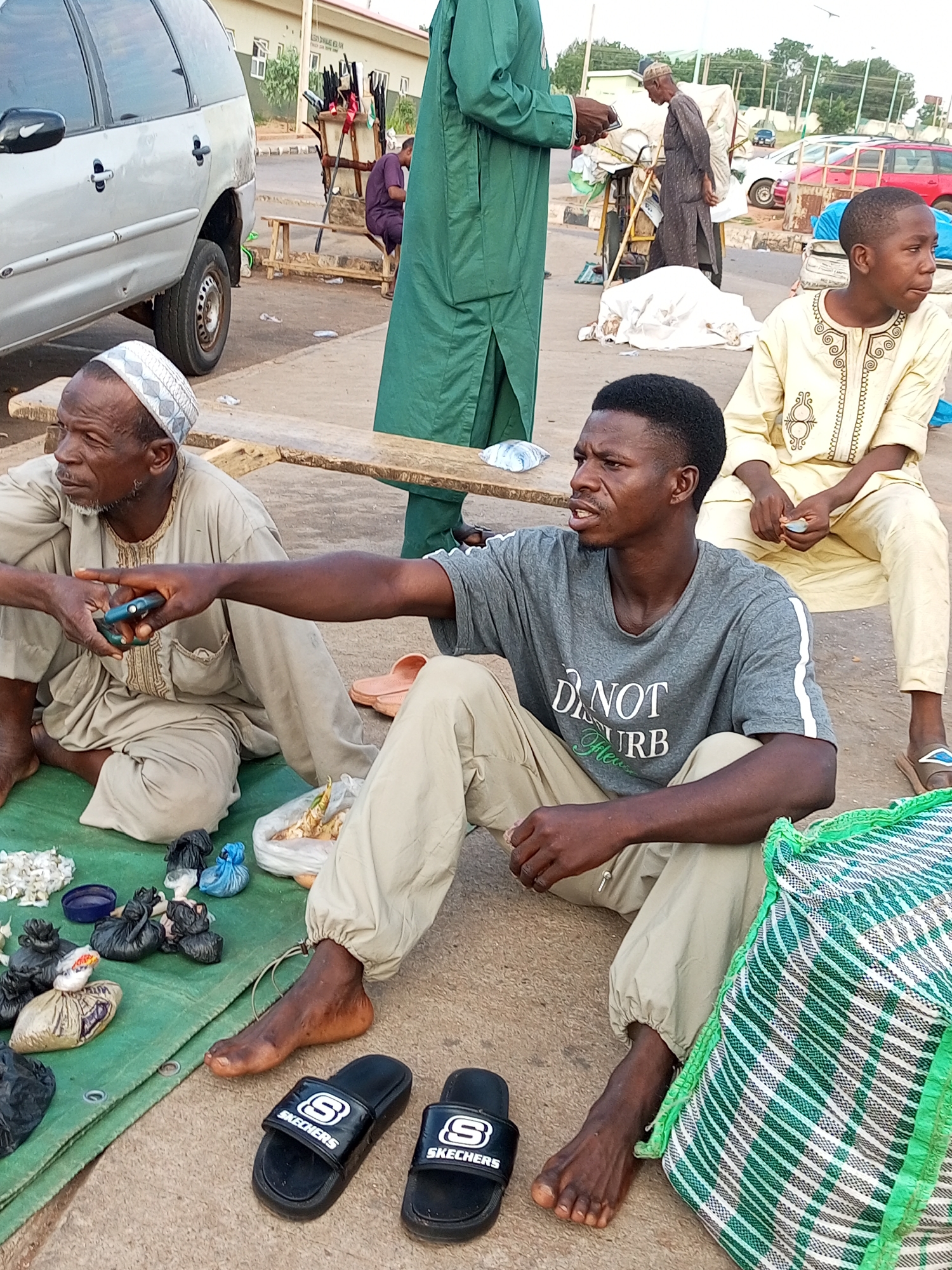 In the city of Gombe, Nigeria, two men are seated on a green mat spread on the ground, engaged in a transaction or discussion. The man on the left, wearing a beige traditional outfit and a patterned cap, holds a blue object, possibly a mobile phone, in his left hand. His right arm is extended, and his hand is gesturing towards the man on the right. The man on the right, younger, wears a grey t-shirt with "DO NOT DISTURB" printed on it and light-colored trousers. He is looking to his left, his mouth slightly open as if speaking.
Various items are displayed on the green mat in front of them. These include small black plastic bags containing what appears to be produce or goods, a blue container, and a larger plastic bag filled with roots or vegetables. To the right of the men, a large green and white striped woven bag rests on the ground. Two black "Skechers" branded flip-flops are placed in front of the man on the right.
In the background, a silver vehicle is parked. Further behind, several individuals are visible, some standing near a market stall with various items displayed. One person, in a long green robe, stands near the vehicle. Another person, in traditional attire, walks past a cart laden with goods. In the distance, cars are parked on a street, and buildings, possibly shops or administrative structures, are visible. The ground surface is paved, suggesting a street or market area.