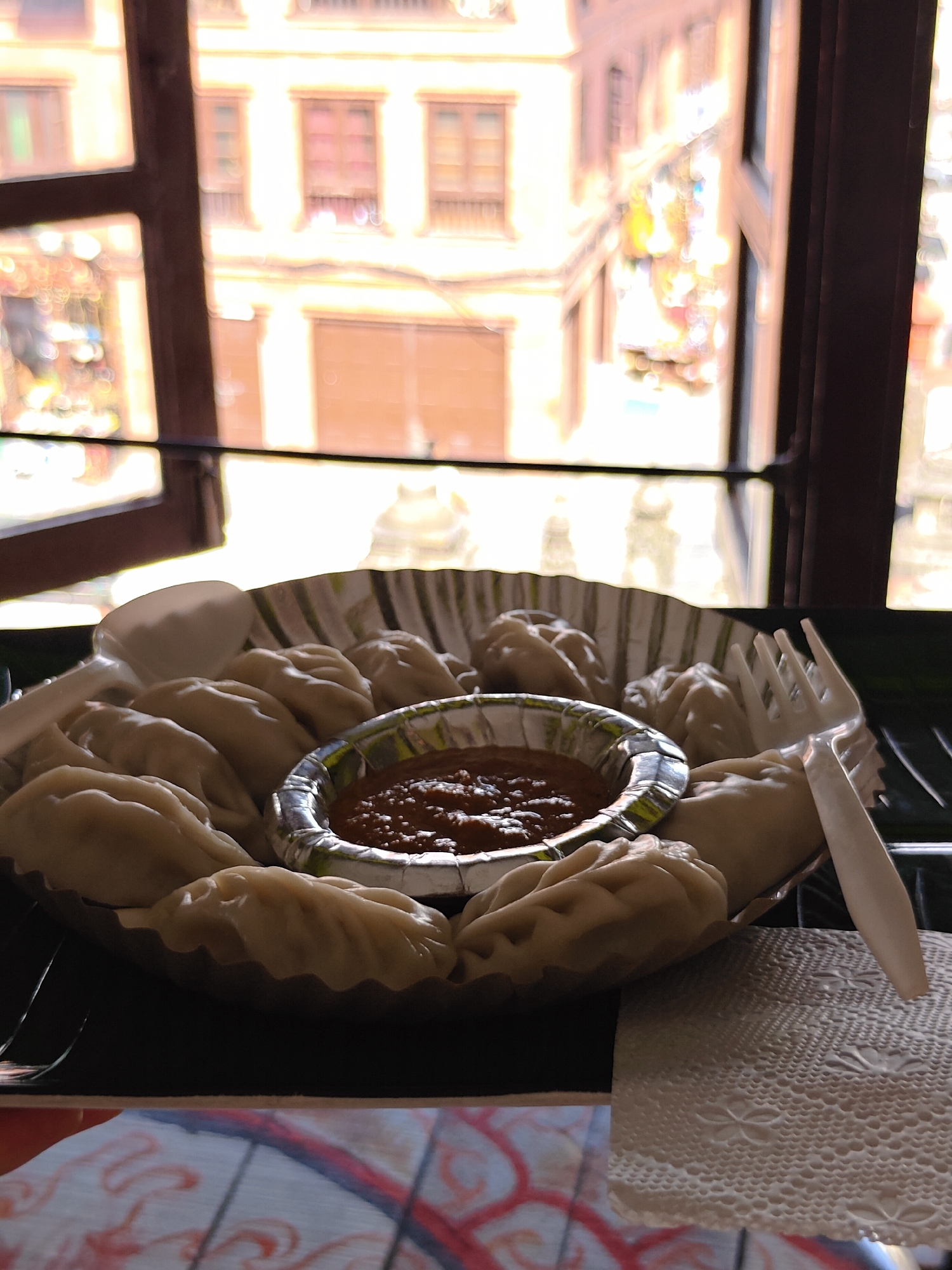 A plate of momos is presented in front of a window with a view of building facades. The momos are arranged in a circular fashion on a paper plate with a corrugated edge, with a small foil container holding a reddish dipping sauce in the center. A plastic spoon and fork rest on either side of the plate. The window shows an urban landscape with multiple stories of buildings, some with windows and balconies, and blurred elements suggesting activity outside. The lighting suggests it is daytime. The scene is likely in Kathmandu Metropolitan City, Nepal, given the presence of momos, a popular dish there.