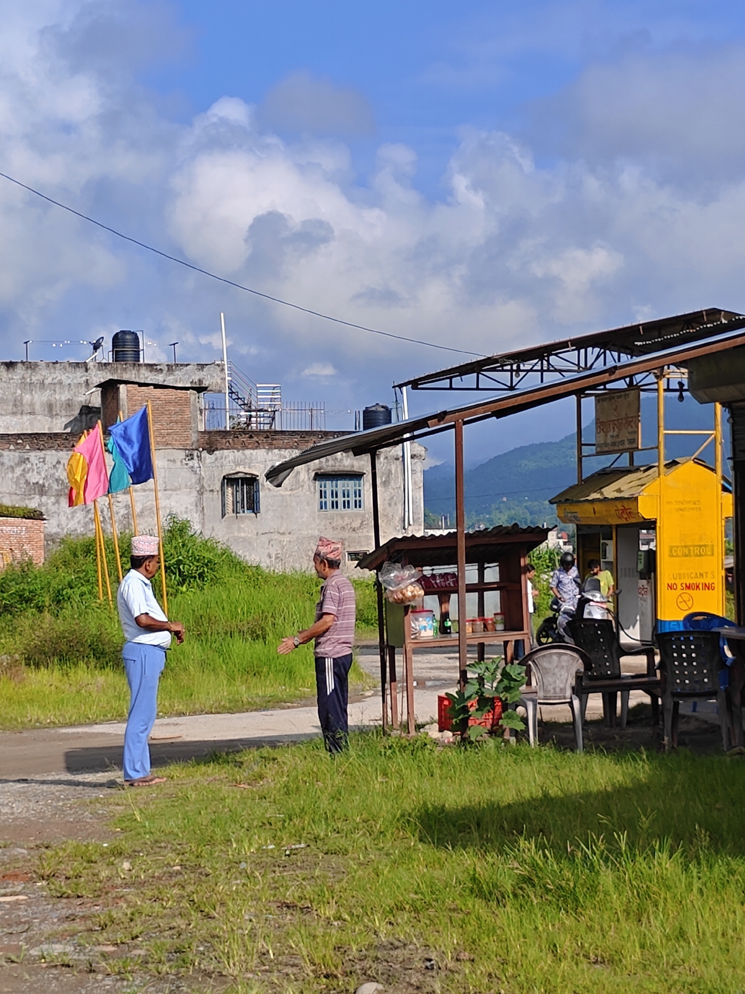 This image depicts an outdoor scene in Tharpu, Nepal, featuring two men standing in front of a structure that appears to be a small roadside establishment or a gas station. The man on the left is wearing a light blue short-sleeved shirt, light blue pants, and a patterned cap. He is standing with his hands clasped in front of him, facing the other man. The second man, on the right, is wearing a striped short-sleeved shirt and dark pants, also with a patterned cap. He is facing the first man with his arms slightly outstretched, as if in conversation.
Behind the men, there are three colorful flags (pink, yellow, and blue) mounted on poles. Further back, a building with a concrete facade, several windows, and a water tank on the roof is visible. The environment is characterized by lush green grass and vegetation surrounding the paved area where the men are standing.
To the right of the men, there is a metal-framed structure with a corrugated roof, likely an awning. Underneath this awning, a yellow structure with signage, possibly a fuel pump or service booth, is present. The signage includes text in a script that appears to be Nepali. Chairs are set up in front of this structure, and a motorcycle is parked nearby. The background shows a hilly landscape with trees under a partly cloudy blue sky. The overall impression is that of a rural or semi-urban setting in Nepal, possibly during daylight hours.
