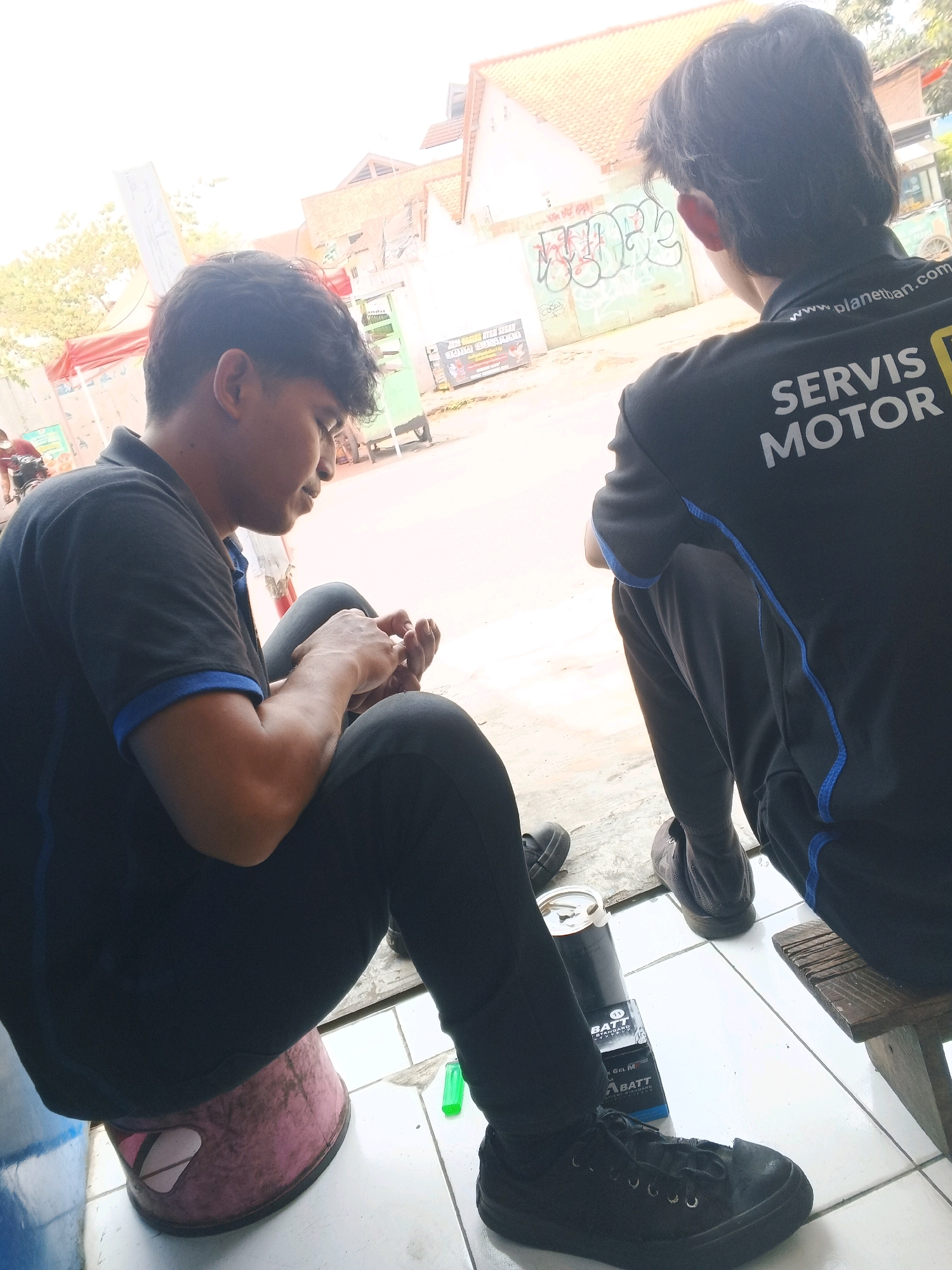 Two young men are sitting outdoors, possibly in a workshop or street-side area in Taman, Indonesia. The man on the left, wearing a black polo shirt with blue trim and black pants, is seated on a pink bucket and appears to be examining something in his hands. The man on the right, facing away from the camera and wearing a black long-sleeved shirt with "SERVIS MOTOR" and "www.planethan.com" printed on the back, is seated on a wooden stool. In the foreground, on the tiled ground, are several objects including a black cylindrical container and a cardboard box labeled "BATT GEL M". In the background, there is a building with orange-tiled roofs and graffiti on a wall, suggesting an urban environment. The lighting suggests it is daytime.