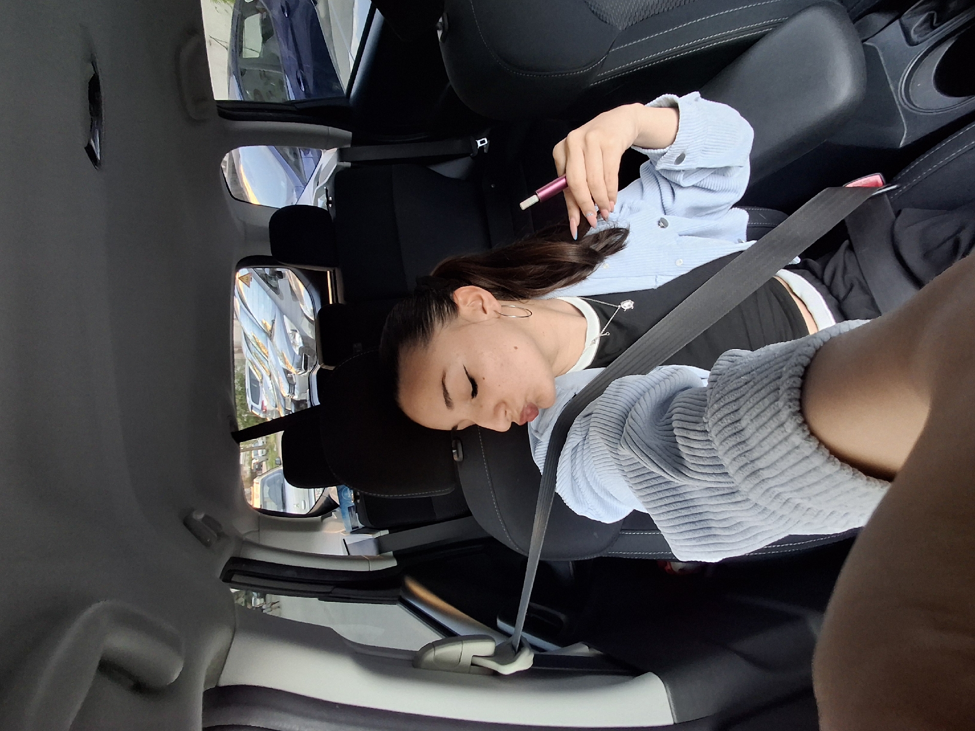 A young woman is depicted resting or sleeping in the back seat of a car, seen from a high-angle, rotated perspective. She has dark hair tied in a ponytail and is wearing a light blue ribbed sweater or cardigan over a black top with a white trim. A silver necklace with a dark pendant and hoop earrings are visible. Her eyes are closed, and her lips are slightly pursed, indicating a state of repose. In her left hand, she holds a pink and white vape pen. A black seatbelt is fastened across her chest. The car's interior features black, textured seats and a light grey ceiling. Through the car windows, bright daylight is visible, along with reflections of other vehicles and buildings, suggesting the car is parked outdoors during the daytime. No visible text is present in the image. The scene conveys a moment of rest or slumber within a vehicle.