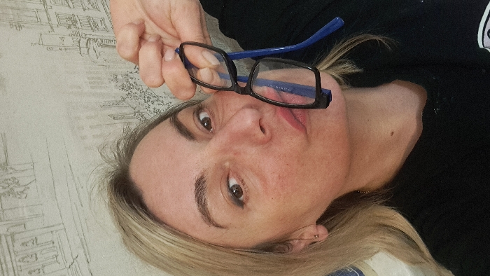 A close-up selfie depicts a blonde female subject indoors in Bel'kovo, Russia. The woman, shown from the chest up, has light-colored hair, dark eyebrows, and eyeliner applied to both eyes. She exhibits some redness on her facial skin. Her lips are pursed forward, and a small, light-colored stud earring is visible in her left earlobe. She wears a dark, possibly black, long-sleeved garment. Her right hand holds a pair of eyeglasses with black frames and blue temples, positioning them in front of her lower face. The background consists of a light-toned wall covered in wallpaper featuring a repeating pattern of sketched monochromatic architectural designs, including buildings and faint tree-like structures.