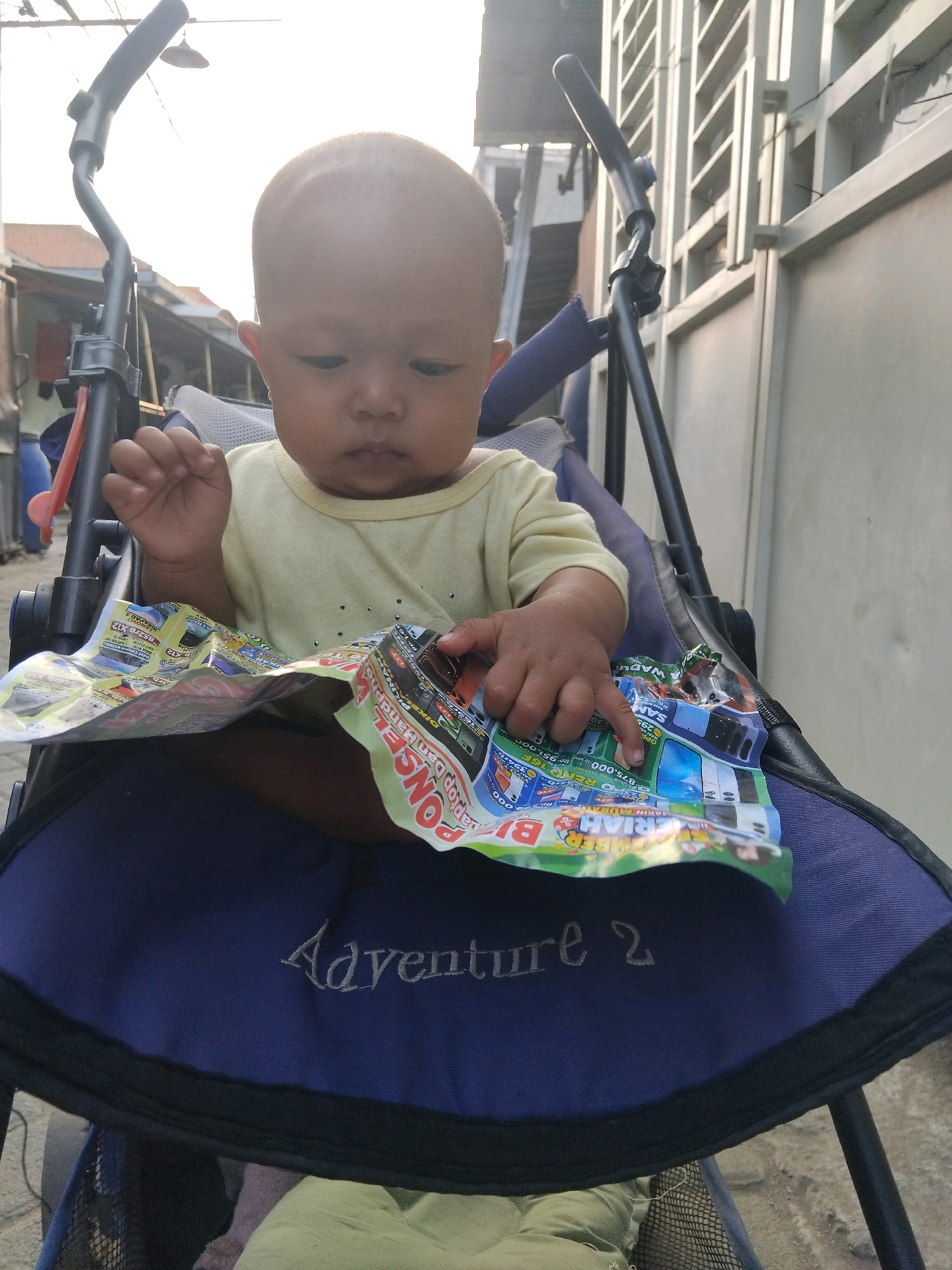 An overhead, slightly downward-facing medium shot shows a bald toddler sitting in a dark blue stroller, holding a colorful, folded flyer. The toddler, wearing a pale yellow t-shirt, looks down at the flyer with their right index finger pointing at it. The stroller has black tubing and a padded dark blue seat with "Adventure 2" embroidered in white thread on the front. The background shows an outdoor scene with a light-colored, textured wall on the right, and a blurry view of buildings and overhead wires on the left, suggesting an urban or suburban setting in Kepuh Kiriman, Indonesia. The sunlight is bright and casts a glow.