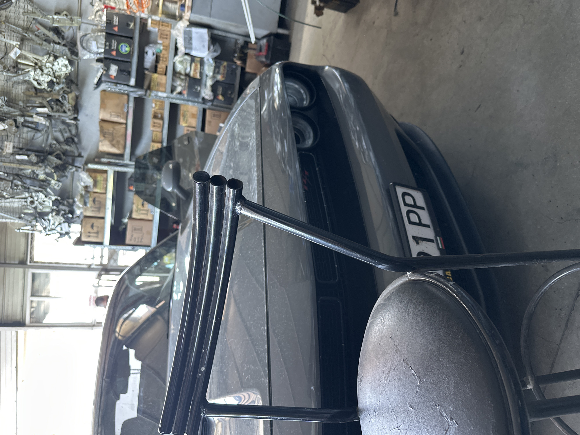 A dark gray Dodge Challenger is parked in a garage, with shelves filled with car parts and boxes in the background. A black metal stool with a worn leather seat is in the foreground, partially obscuring the car's hood and front bumper. The car's headlights and part of its grille are visible. The license plate on the car reads "91 PP". The garage appears to be dimly lit, with bright light coming from windows or doors at the far end.