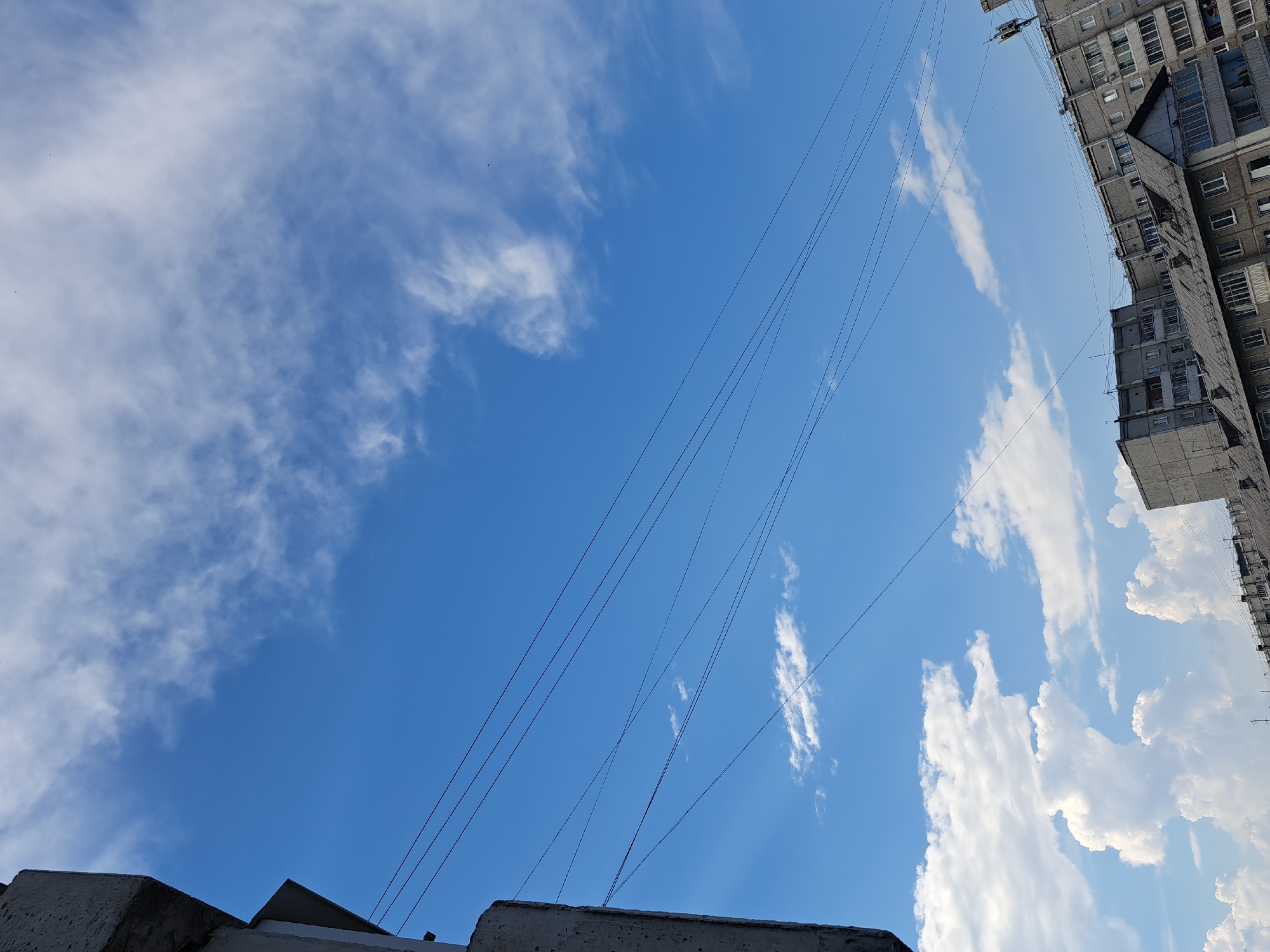 The image shows a view of the sky and buildings in the city of Irkutsk, Russia. The sky is bright blue with scattered white clouds. Several power lines stretch across the frame. In the lower portion of the image, several apartment buildings are visible, characterized by their concrete construction and numerous windows. Some of the buildings have pitched roofs with chimneys. The perspective suggests the photo was taken from a higher vantage point, looking upwards. There are no people visible in the frame.
