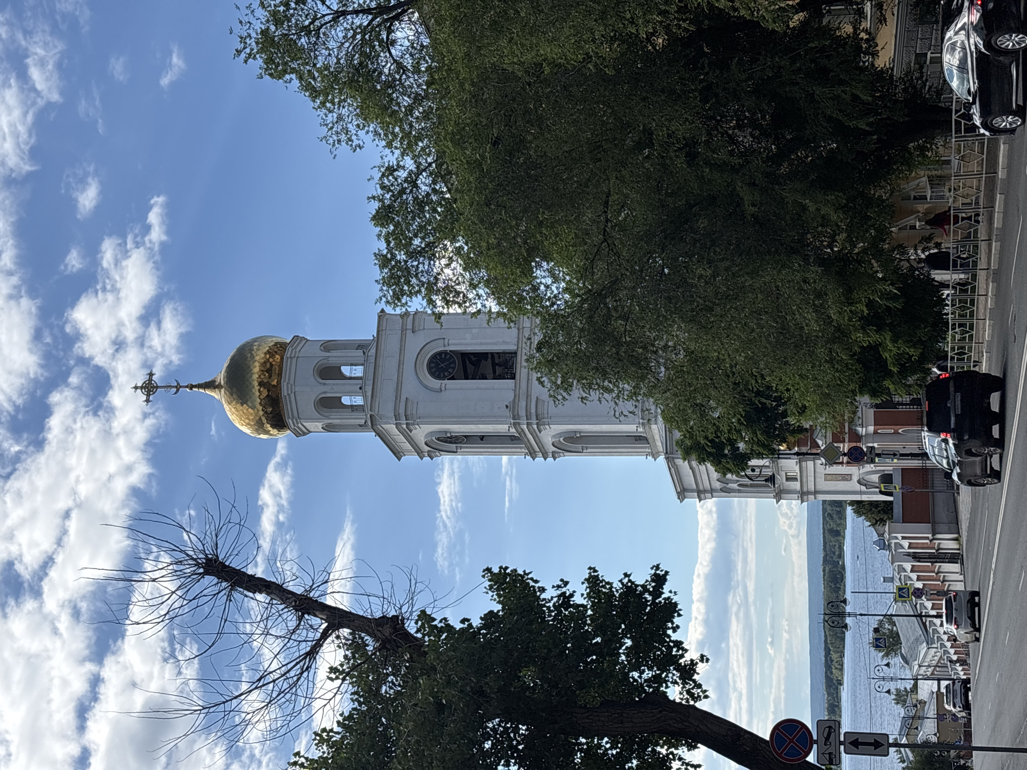 The image shows a view of the Transfiguration Cathedral in Novokuybyshevsk, Russia. The cathedral's white bell tower with a golden dome and cross is prominent against a blue sky with scattered white clouds. A large, bare tree branch extends from the left side of the frame, obscuring part of the sky and the upper portion of the bell tower. Lush green trees frame the right side of the bell tower and obscure some of the lower parts of the cathedral. In the foreground, a street slopes downwards towards a body of water, likely a river or reservoir, with a distant treeline on the opposite bank. Several cars are visible on the street, including a black SUV in the right lane and white cars further down the road. Traffic signs are present on the left side of the street. A few pedestrians are visible on a sidewalk behind a decorative fence to the right of the street. The architecture of the buildings along the street suggests a mix of older and more modern styles. The overall impression is a sunny day in an urban setting with a prominent religious landmark.