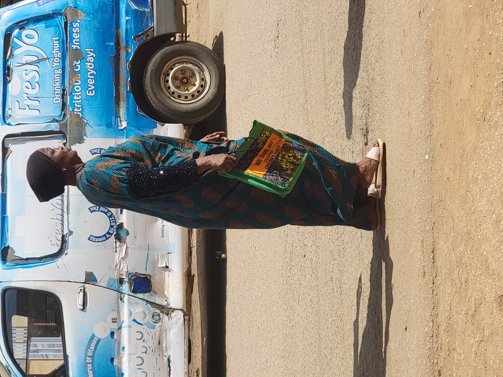 The image, taken in Maiburiji, Nigeria, captures a sunlit outdoor scene featuring a woman standing on a dusty roadside. The woman, viewed in profile, is wearing a long, flowing dress with a teal and brown patterned design and a dark head covering. She has light-colored flip-flops on her feet. In her right hand, she holds a vibrant green bag.
To her left is a blue van, prominently branded with "FreshYo" in large white lettering. Additional text on the van includes "Drinking Yoghurt" and "Nutrition Goodness, Everyday!". Further details on the van's exterior mention it as a "Source Of Vitamins" and containing "Vitamin A, B6, B12, D and Zinc." The branding on the van appears somewhat worn and peeling in various areas. A spare tire is visible towards the rear of the van.
The green bag the woman is holding displays clear text: "DATE: 13 APRIL, 2024", "COURTESY", and "GROOM'S FAMILY." The bright sunlight casts distinct, long shadows on the ground, suggesting it is midday or early afternoon with clear weather. The woman appears to be standing or waiting by the roadside.