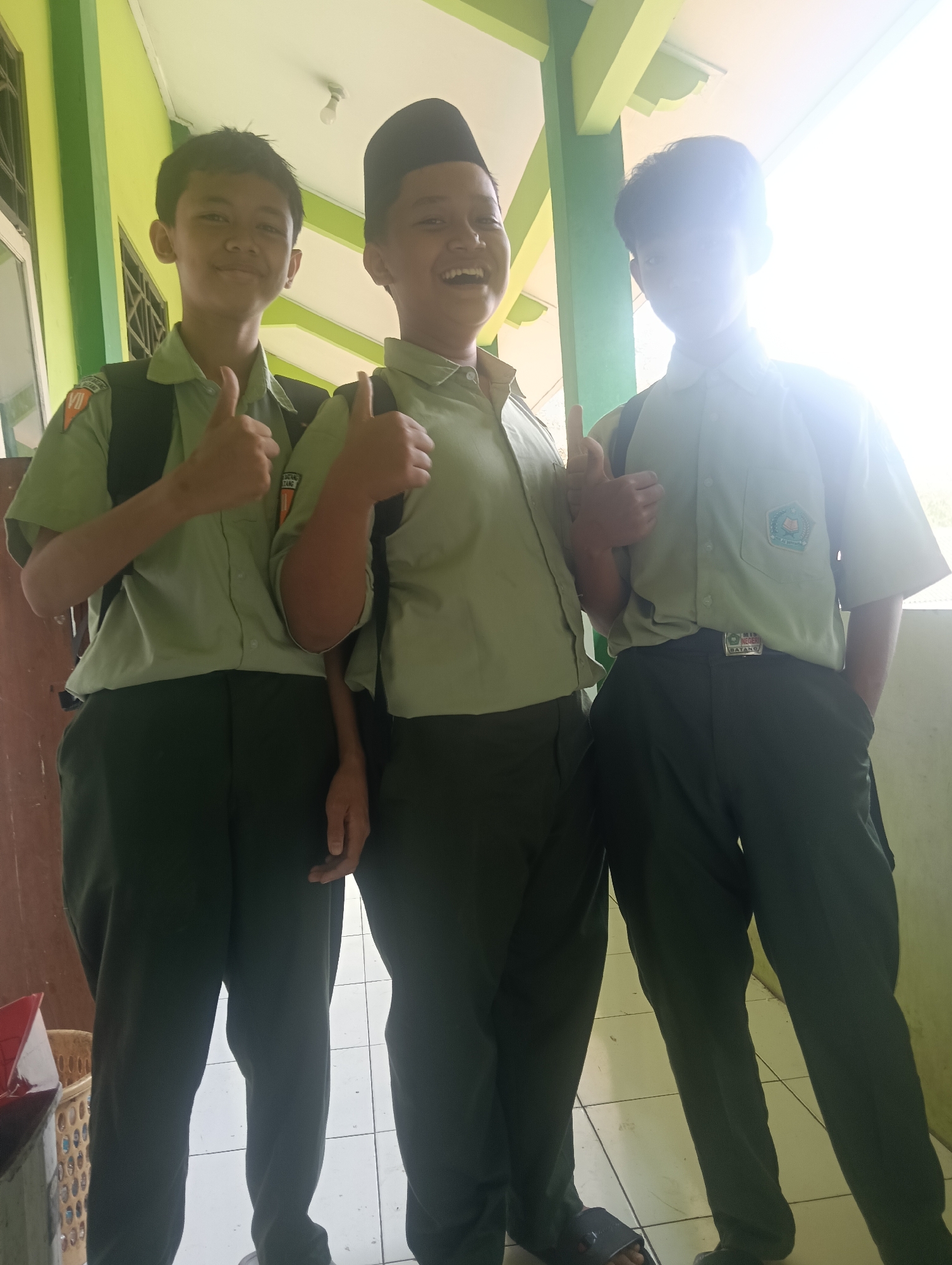 Three young males, appearing to be of Indonesian ethnicity and wearing school uniforms consisting of light green shirts and dark pants, stand in front of a building with green trim. The male on the left has short dark hair and gives a thumbs-up. The middle male wears a black peci cap and is laughing, also giving a thumbs-up. The male on the right, facing away from the camera and backlit by bright sunlight, has dark hair and is holding his hand in his pocket, giving a thumbs-up. They all appear to be wearing backpacks. The environment suggests an outdoor covered walkway or veranda, with tiled flooring and a yellow wall. Based on the context of school uniforms and common building structures in Indonesia, the location is likely an educational institution in Adinuso, Indonesia.