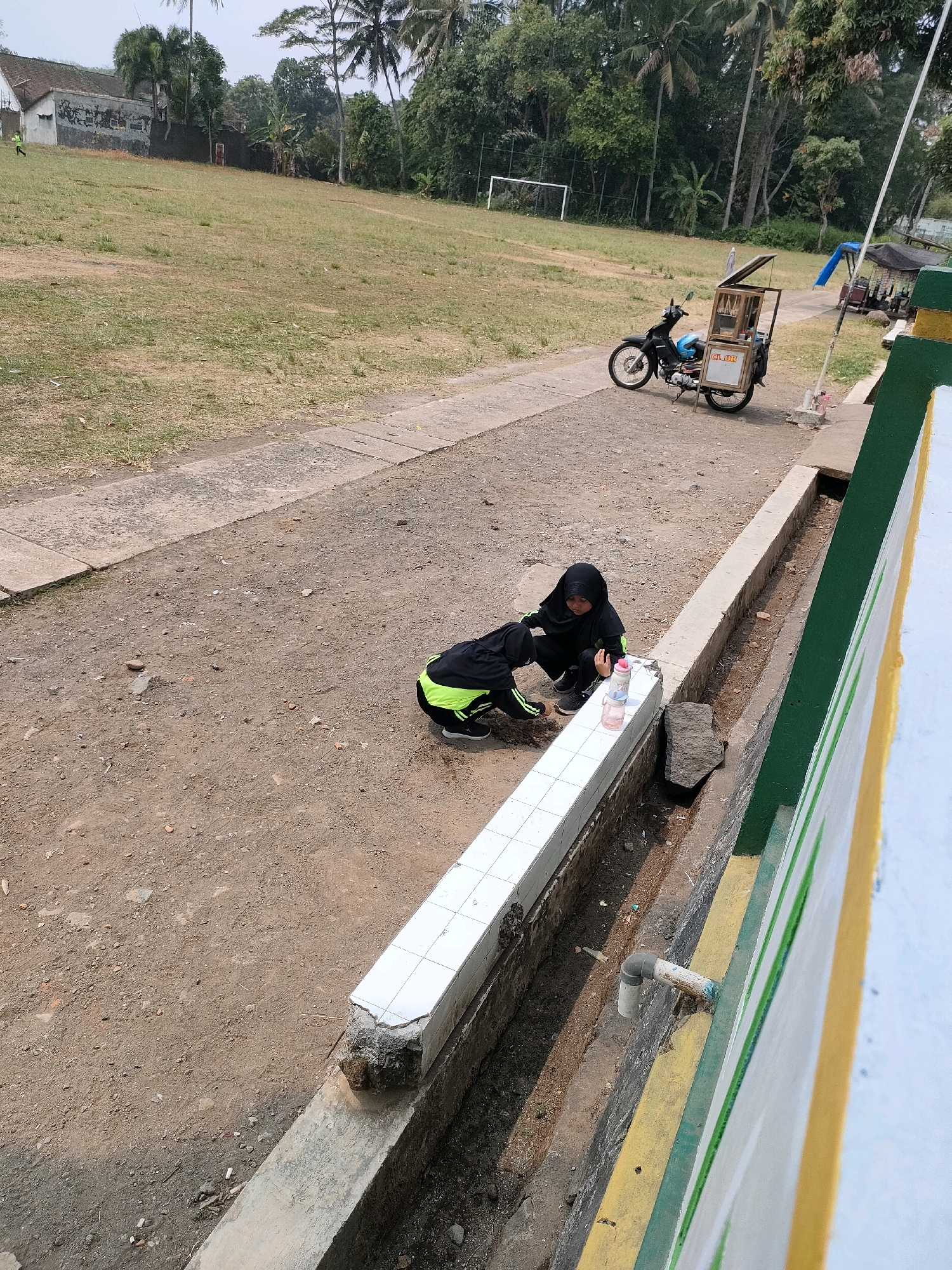 The image depicts an outdoor scene in Bojong, Indonesia. Two individuals wearing dark athletic attire with lime green accents are crouched on a dirt path next to a white tiled wall. One person is reaching towards a water bottle. Behind them, a dirt field with patches of dry grass stretches towards a building and trees. A motorcycle with a cargo carrier is parked on a concrete path to the right of the field. A soccer goal is visible in the distance on the field. The environment appears to be a rural or semi-rural area.