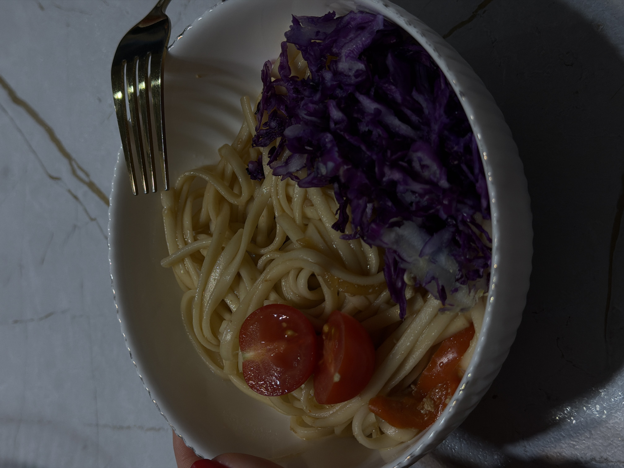 An overhead, close-up shot captures a meal served in a white bowl, held by a hand with red nail polish at the bottom left. The bowl contains light-colored pasta, likely linguine or fettuccine, topped with a generous portion of vibrant shredded purple cabbage and two halved red cherry tomatoes. A third piece of red vegetable, possibly another tomato or a red pepper, is partially visible near the bottom right. A golden-toned fork rests on the rim of the bowl, with its prongs extending towards the pasta.
The meal is presented on a light-colored surface, which appears to be a marble or veined countertop or table. The lighting is dim and directional, creating strong shadows across the scene and casting a reflective sheen on the fork and the surface of the food. The overall ambiance suggests an indoor setting, possibly during the evening or in a low-light environment. There are no visible location cues specific to Chaikovsky, Russia, nor any discernible text or explicit emotions. The image portrays a simple, colorful meal prepared and ready for consumption.