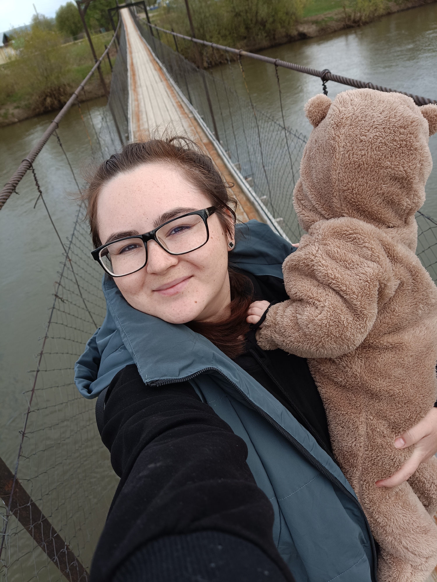 A woman wearing glasses and a blue jacket is holding a baby in a brown bear costume. They are standing on a suspension bridge. A river flows below the bridge. The location is in Posad, Russia.