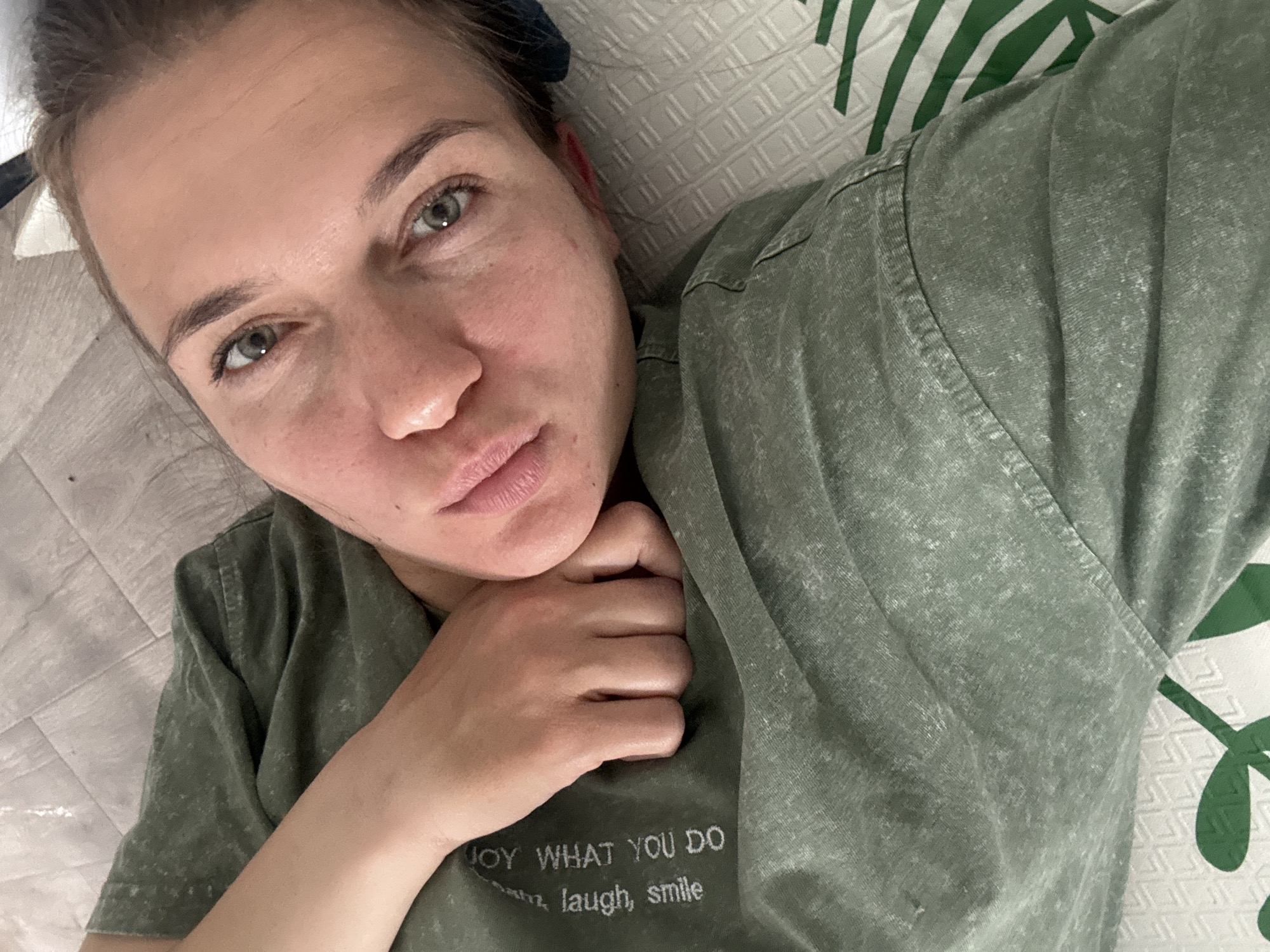 The image features a woman lying down in an indoor setting, looking directly at the camera in what appears to be a selfie or close-up portrait. She is positioned diagonally across the frame, with her head towards the upper left.
The woman has light green or hazel eyes and a calm, neutral expression, with her lips slightly pursed. Her hair is pulled back, and a dark hair tie or scrunchie is visible near her left ear. Her complexion appears natural, showing some minor blemishes and freckles.
She is wearing a short-sleeved, olive green t-shirt that has a faded, mottled texture. Her right hand is visible, resting on her chest. Embroidered in white on her shirt, near her hand, is the text: "JOY WHAT YOU DO" clearly visible, followed by "laugh, smile" on the line below. The word before "laugh" is partially obscured.
The background consists of light-colored bedding or a textured surface to her left, and to her right and behind her head, a white pillow or duvet with a bold green geometric pattern, possibly depicting abstract leaves or foliage. The scene suggests a relaxed atmosphere, likely in a bedroom or living space. The time of day and weather are not discernible.
