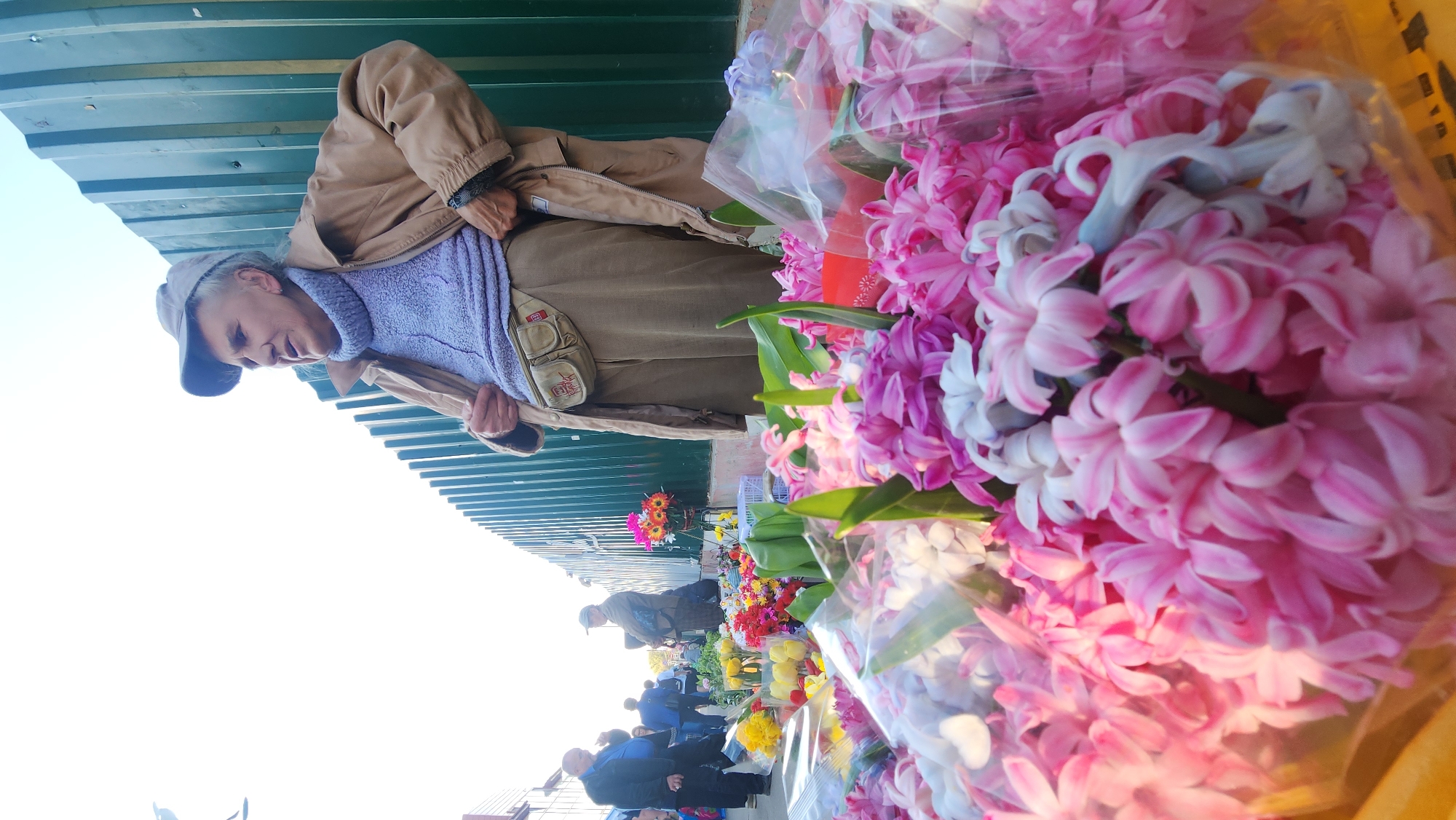 An elderly woman in a brown jacket and hat stands at a flower stand. The stand is filled with colorful flowers including pink hyacinths. Other people are in the background. The location is Khimki, Russia.