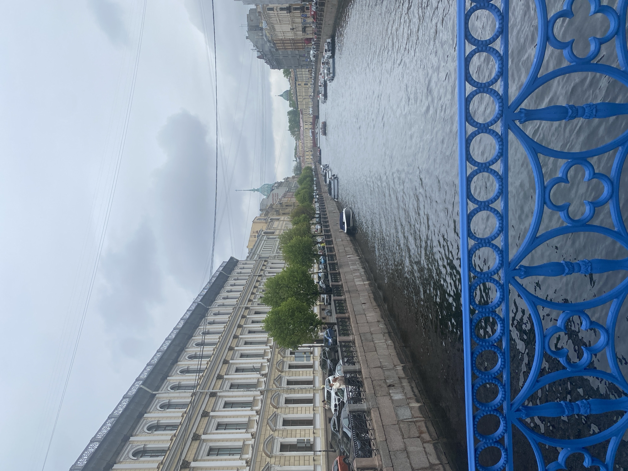 The image depicts a view from a bridge in Saint Petersburg, Russia, likely during overcast weather. The foreground features a section of the bridge's blue metal railing with intricate circular and clover-like patterns. The scene extends into the distance, showcasing a canal or river with numerous boats. Along the waterway's edge, buildings are situated with light-colored facades and ornate architectural details, some lined with trees. Parked cars are visible along the embankment. Power lines stretch across the sky, adding to the urban landscape.