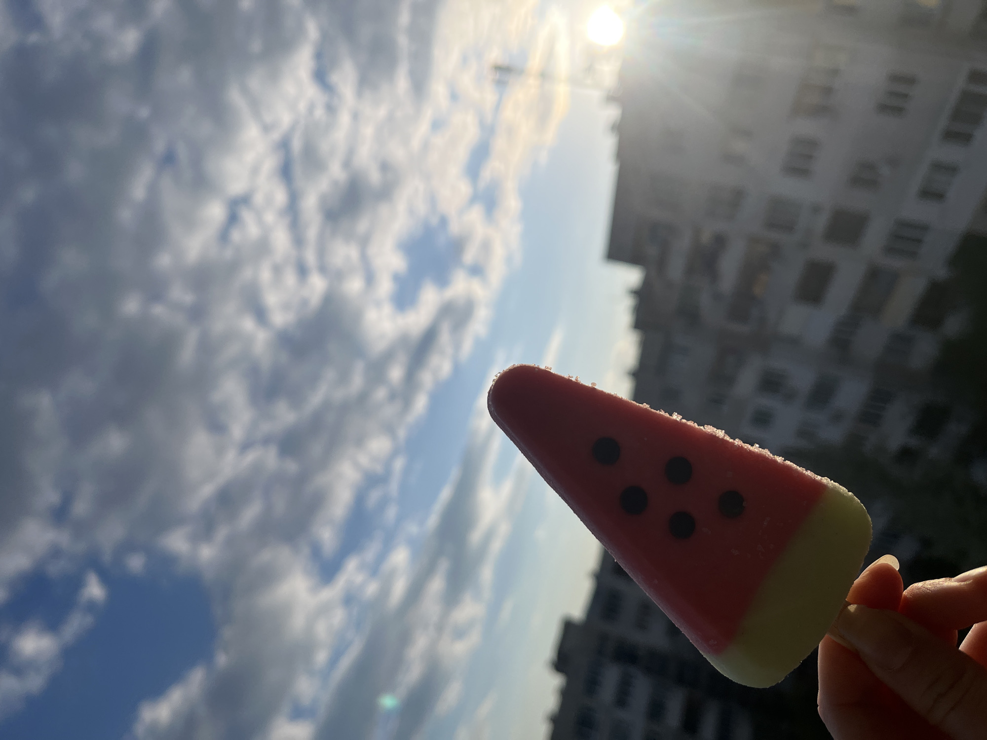 The image presents a vibrant, sun-drenched outdoor scene. In the foreground, a person's hand, with slender fingers and manicured nails, holds a watermelon-shaped popsicle by its wooden stick. The popsicle is distinctively designed, featuring a bright red upper section with four small black 'seeds,' transitioning to a pale yellow-green layer at the bottom, resembling a watermelon slice. Fine ice crystals are visible on its surface, indicating its coldness.
The background is dominated by a bright blue sky with fluffy white clouds, suggesting a clear and pleasant day. A powerful sun, positioned in the upper right quadrant, casts a brilliant lens flare and backlights the entire scene, creating a strong contrast. Partially visible in the distant background, a tall, multi-story urban building, likely an apartment or office block, appears silhouetted and somewhat blurred against the bright sky due to the backlighting and upward angle of the shot.
The activity depicted is the enjoyment of a refreshing cold treat outdoors, evoking feelings of summer, relaxation, and warmth. The time of day appears to be mid-day or afternoon given the high and intense sun. The setting is an urban outdoor environment in Sofia, Bulgaria. No visible text is present in the image.