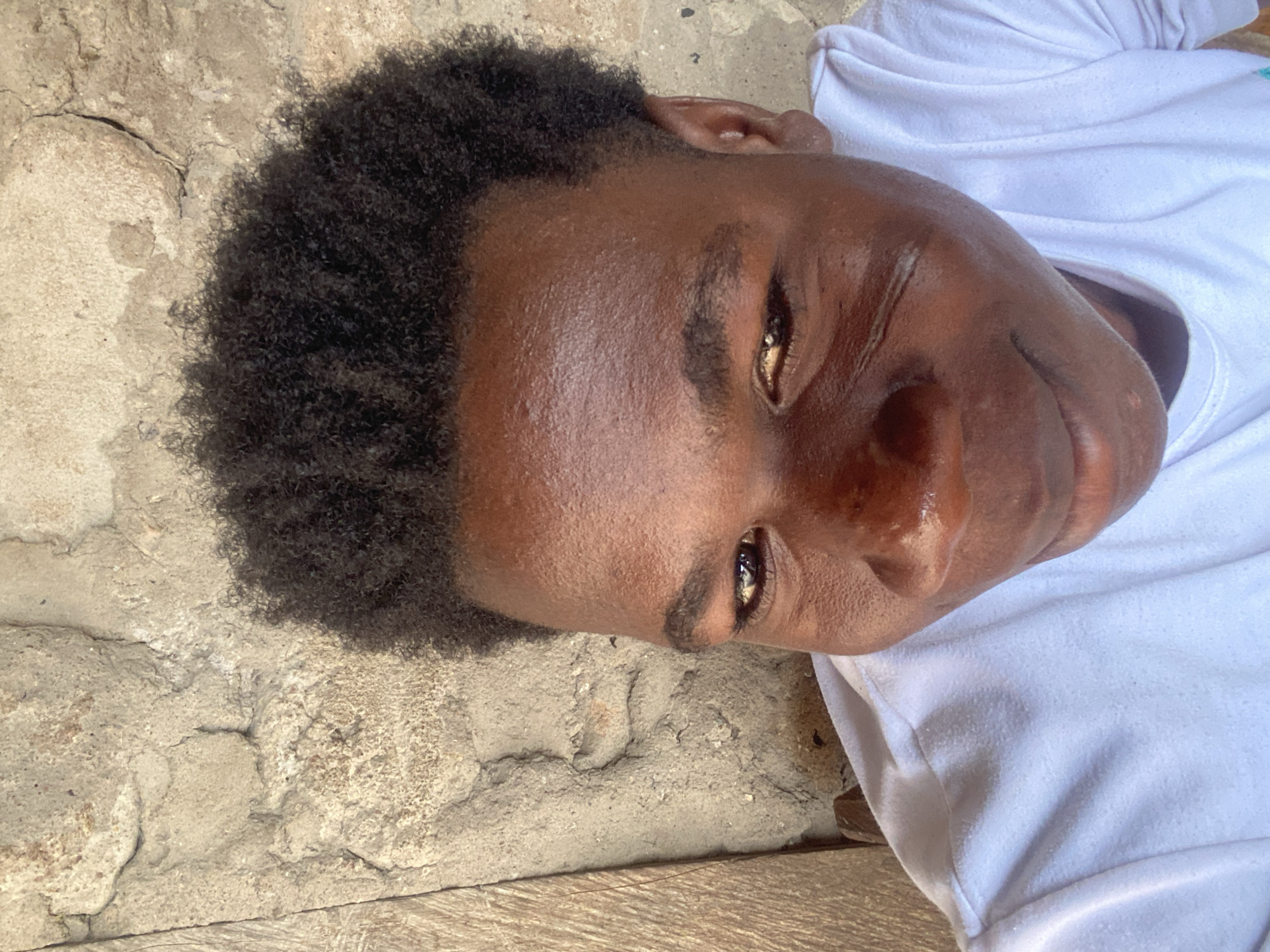 A young, dark-skinned man with short, dark, kinky hair lies sideways, wearing a white shirt. Part of his head rests on a rough, light-colored surface, possibly concrete, with visible cracks and uneven texture. The picture was possibly taken in Mararaba, Nigeria but without additional context, location cannot be confirmed.