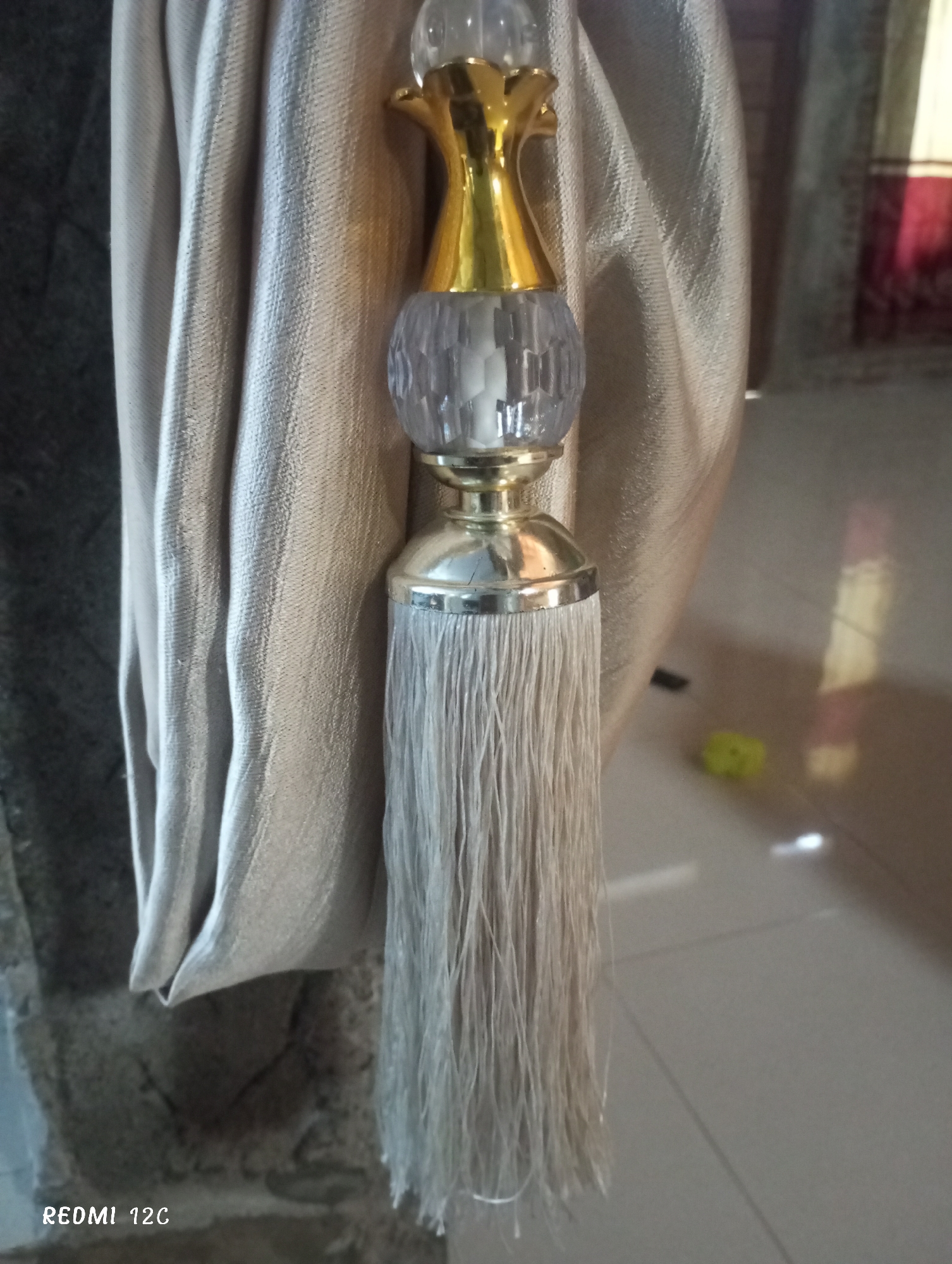 A close-up indoor shot depicts a section of draped, light-colored fabric, possibly a curtain, held back by a decorative tassel. The tassel has a bulbous, multi-faceted clear element, a gold-colored floral top, and a silver-colored base with a long, fine fringe. The fabric is draped against a rough, dark-colored surface, possibly a wall. The foreground floor is light-colored. A yellow object is visible on the floor in the background. A faint reflection is visible on the floor, showing a door with a red section. The words "REDMI 12C" are visible in the lower left corner of the image. The location is Lhokseumawe, Indonesia.
