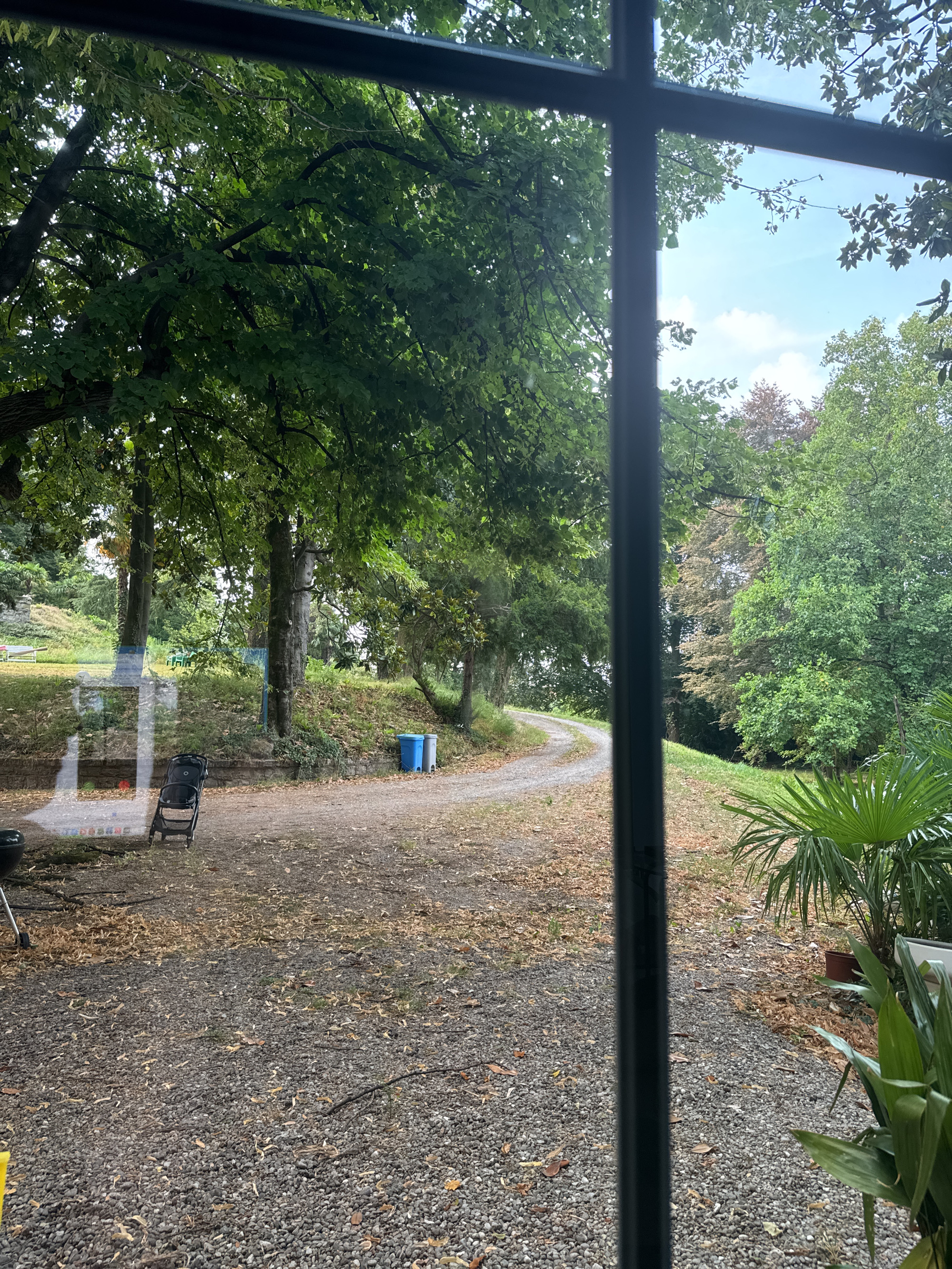 The image shows a view through a window of a winding gravel path in a park-like setting. The path is lined with trees and grass, and there are fallen leaves on the ground. On the left side of the path, there is a charcoal grill and a black folding chair. Further up the path, there are two trash bins, one blue and one gray. In the background, there are more trees and a glimpse of a cloudy blue sky. The scene is likely in the city of Erba, Italy, given the context provided.