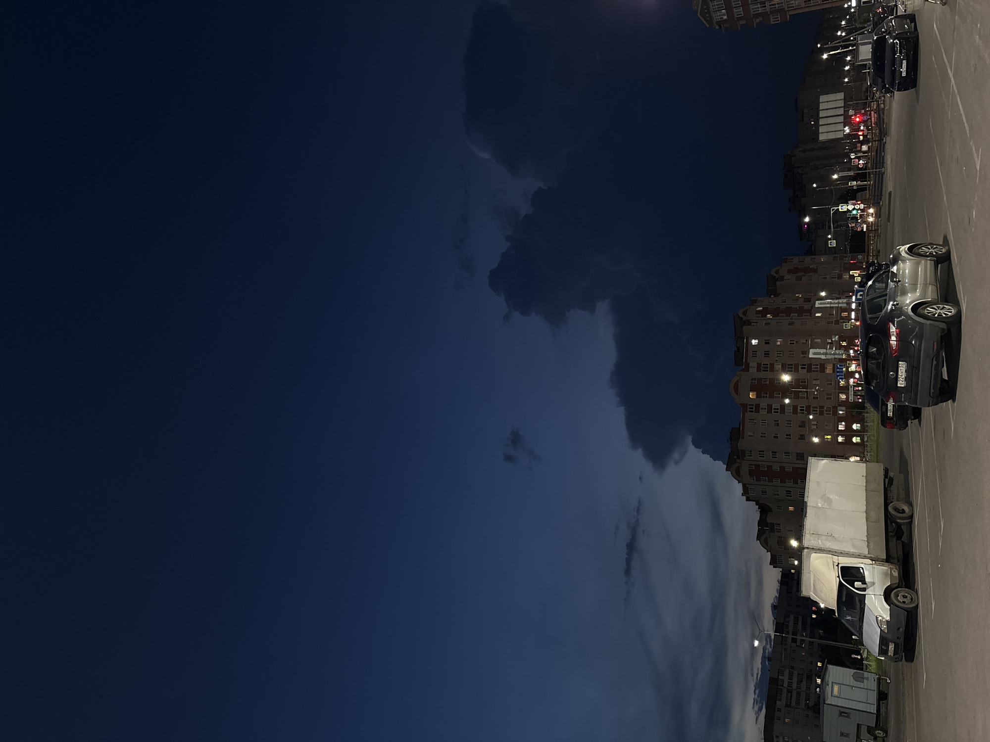 The image depicts a nighttime scene in Veliky Novgorod, Russia. The sky is dark blue with heavy, ominous clouds dominating the upper portion of the frame. In the midground, several multi-story apartment buildings are visible, their windows illuminated, suggesting an urban residential area. Streetlights and traffic signals are lit, indicating the presence of an active roadway or intersection. In the foreground, there are parked vehicles, including a white utility truck with a large cargo box, a dark-colored SUV, and another dark-colored car. A light-colored trailer is parked to the left of the utility truck. The ground appears to be asphalt, likely a parking lot or road surface. The overall atmosphere is dark and somewhat brooding due to the dramatic cloud formations and the time of day.