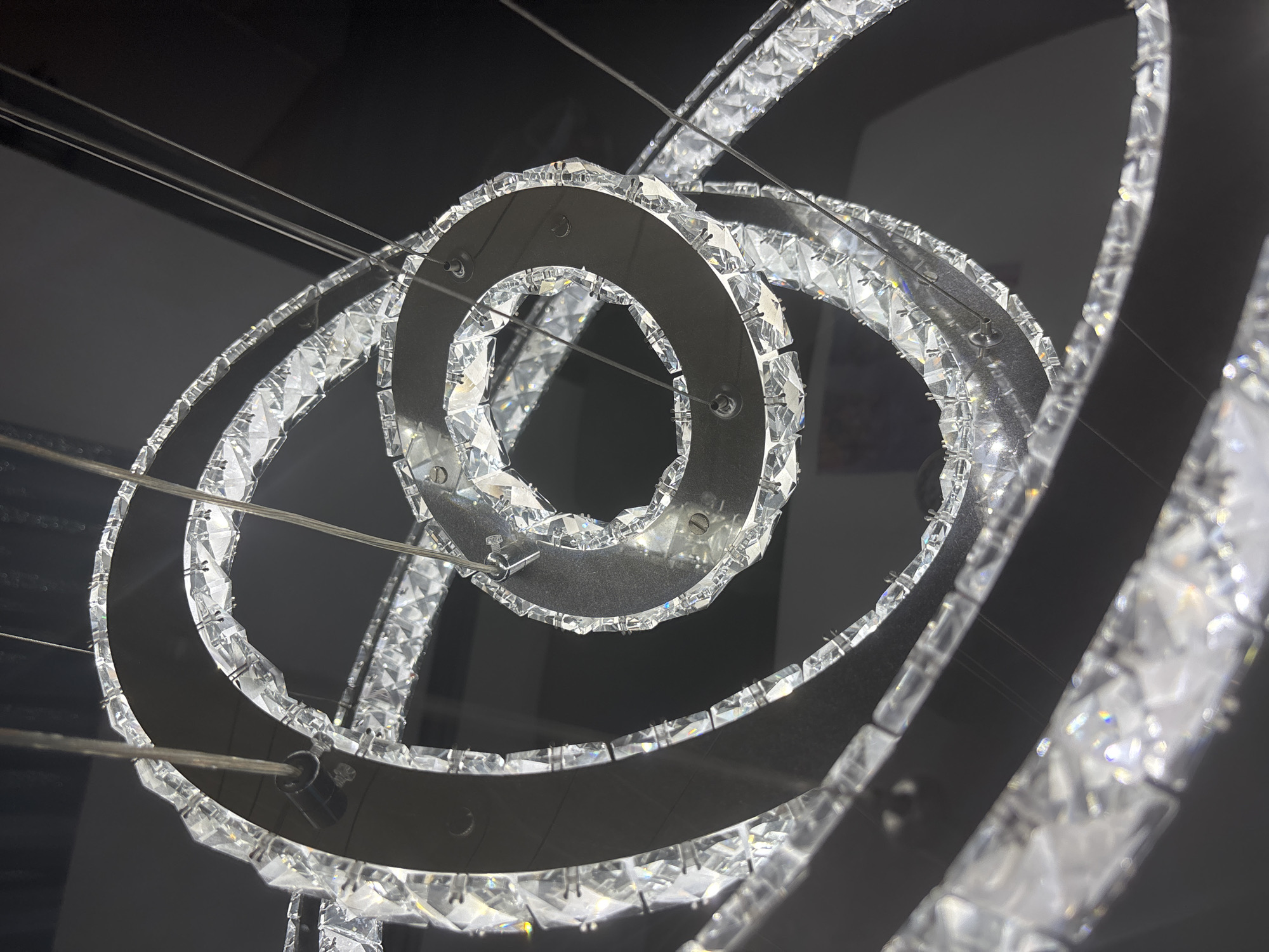 A close-up, low-angle shot captures a modern, circular chandelier adorned with numerous crystal-like elements. The chandelier is suspended by thin wires, and the intricate design features concentric rings, some of which are partially obscured by others. The crystal elements are faceted, catching and reflecting light, creating a dazzling and sparkling effect. The background is dark and out of focus, emphasizing the illuminated chandelier. The overall impression is one of luxury and sophistication. The location is Unknown, Unknown.