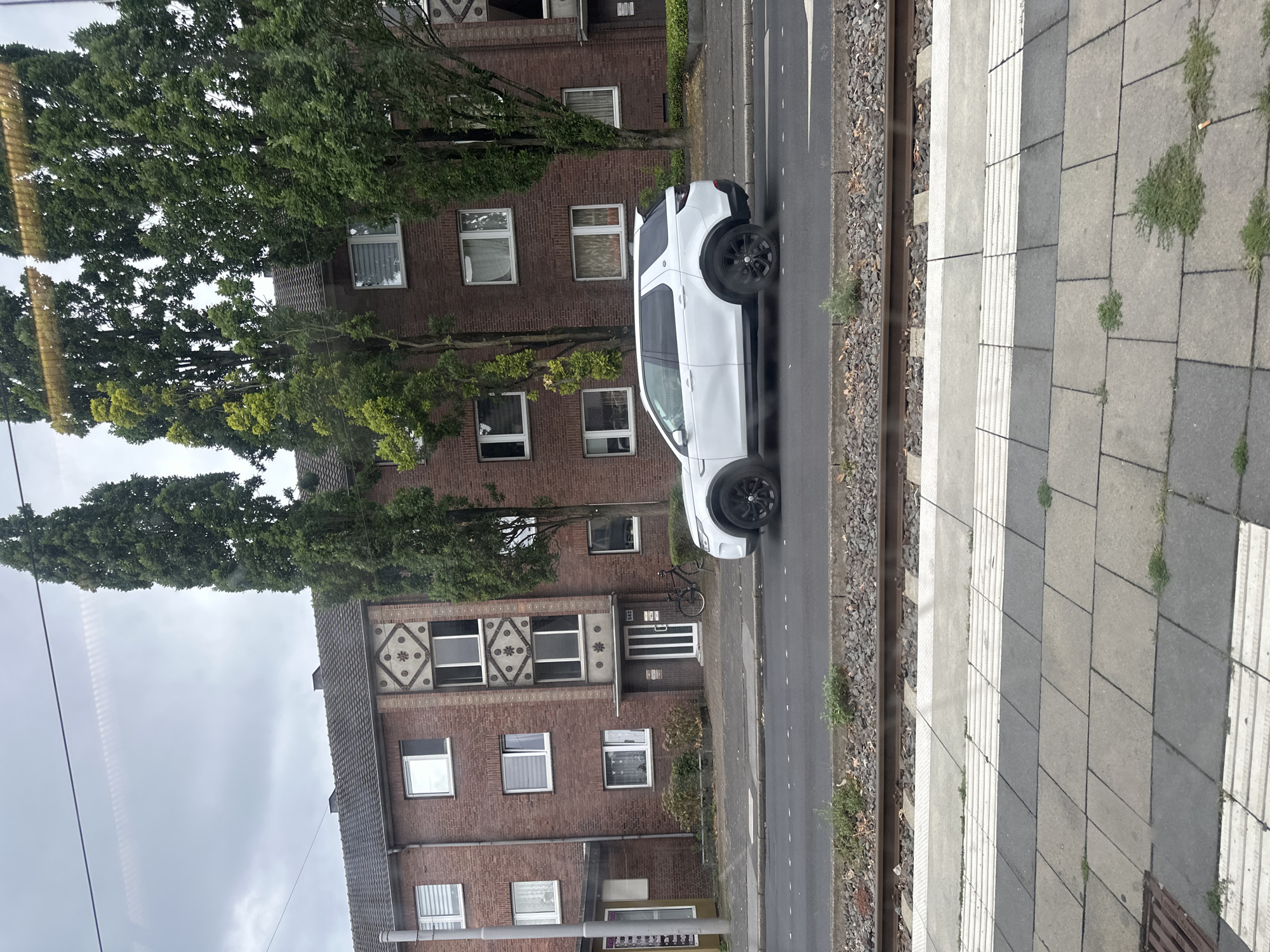 A white SUV is parked on the side of a road in front of a brick apartment building in Krefeld, Germany. The building has several windows and a decorative facade. Trees line the street in front of the building. Train tracks are visible in the foreground, and a platform is to the right of the tracks. The sky is overcast.