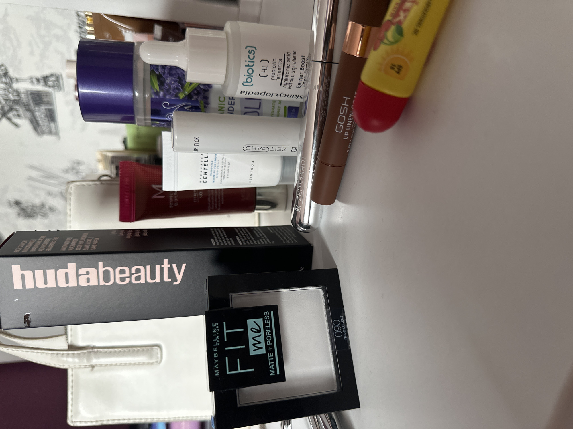 The image shows a collection of cosmetic products arranged on a white surface. The brands represented include Huda Beauty, Maybelline, Skincyclopedia, Skin1004, GOSH and some items without clear labels. The Huda Beauty product is in a black box with pink lettering. A Maybelline Fit Me powder compact is visible in a black case with the shade 090 Translucent. Several skincare items are present, including one labelled "bioticos" from Skincyclopedia, as well as products from Skin1004. A GOSH Lip Liner is also visible. A purse is located on the left of the image.
There is no information available in the image to indicate the city Djerman in Bulgaria or the exact location, scene, or details regarding any people.