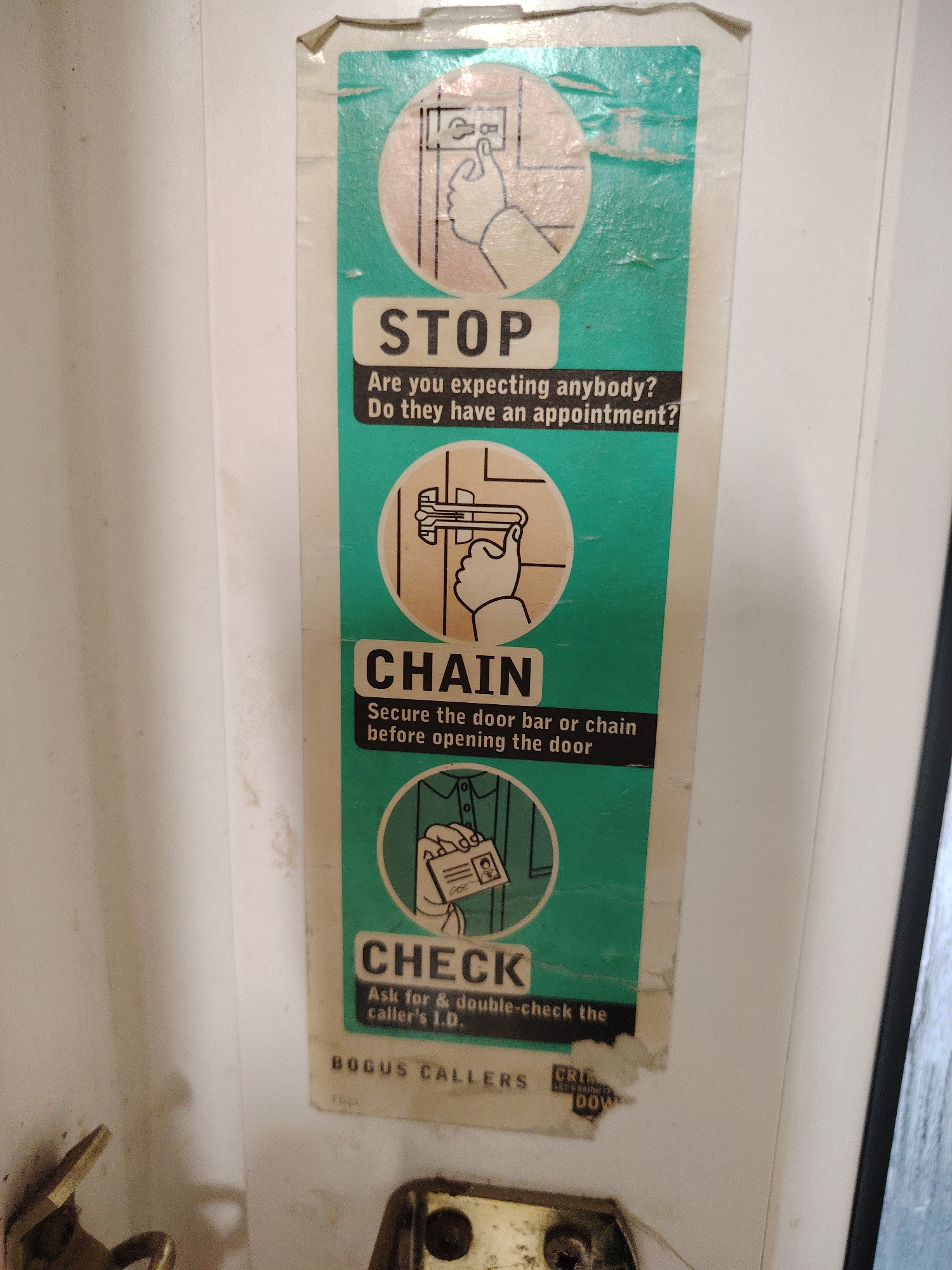An aged safety poster is affixed to the inside of a white or off-white door, likely in a residential setting in Huntingdonshire, United Kingdom. The poster, predominantly faded teal or green with white and black text, shows signs of wear, including peeling edges, creases, and discoloration.
The poster provides three steps for door safety, each accompanied by an illustrative circular graphic:
1. **STOP**: The top section features an illustration of a hand pressing a doorbell. The accompanying text reads, "STOP / Are you expecting anybody? Do they have an appointment?"
2. **CHAIN**: The middle section depicts a hand securing a door chain or bar. The text advises, "CHAIN / Secure the door bar or chain before opening the door."
3. **CHECK**: The bottom section illustrates a hand holding an identification card. The text instructs, "CHECK / Ask for & double-check the caller's I.D."
At the very bottom of the poster, partial text is visible: "BOGUS CALLERS" and fragmented words like "CRI" and "DOW," likely part of a crime prevention message.
Below the poster, parts of metallic door hardware are visible, including a golden-colored plate with screws, possibly part of a lock or latch mechanism, and another brass-toned component on the lower left. The overall scene suggests a focus on home security and awareness against unsolicited or fraudulent callers.