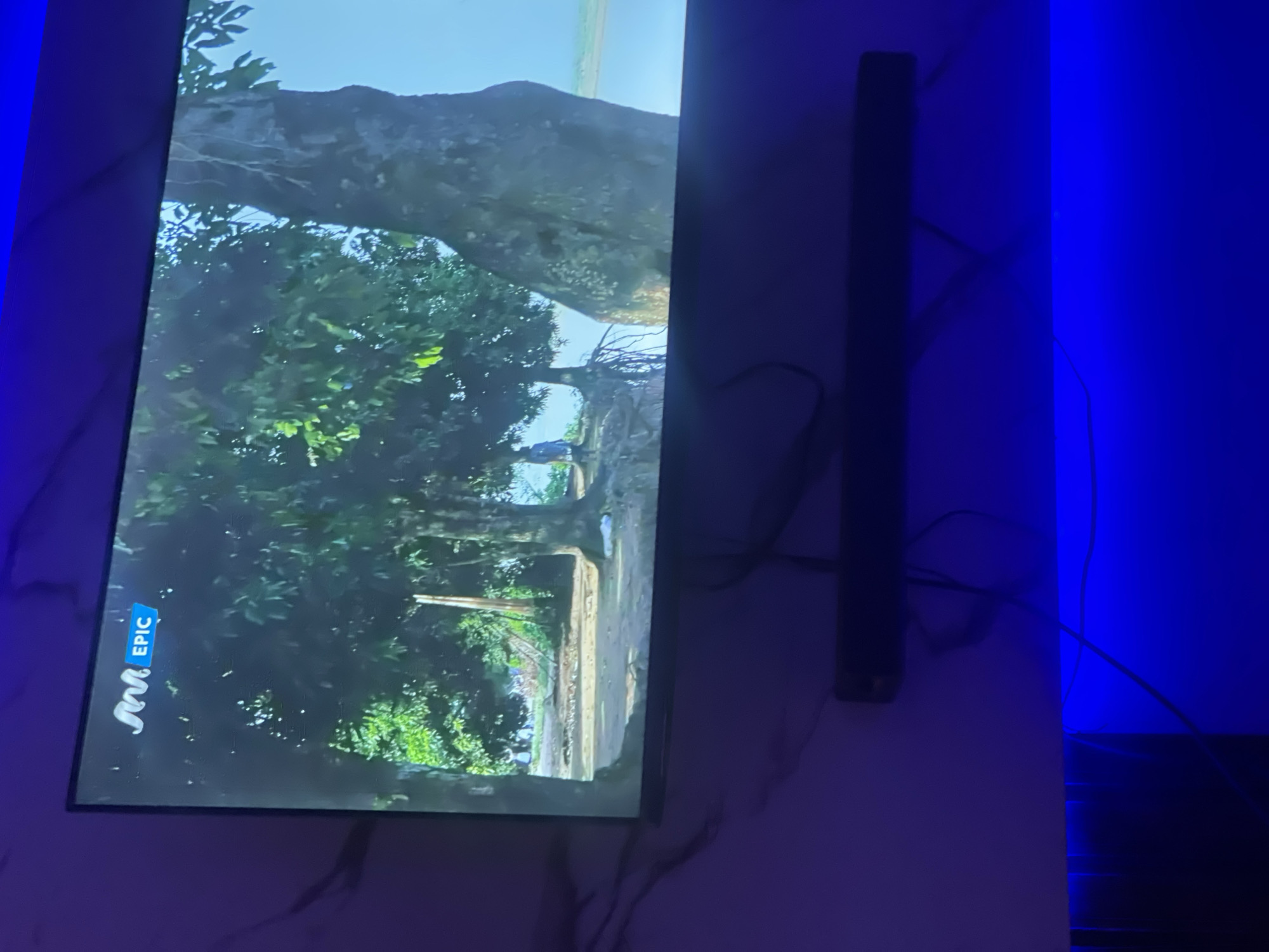 The image shows a television mounted on a wall with a scene of a forest being displayed. The television has the "EPIC" logo on the bottom left corner. To the right of the TV, there is a tall black object, possibly a speaker or soundbar. The wall behind the TV has a marble-like pattern. The surrounding environment is illuminated with blue light. The specific location within Lokoja, Nigeria, cannot be determined from the visual information provided.