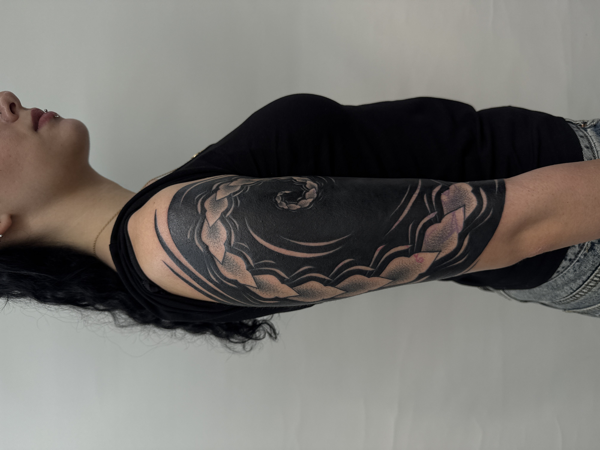 A sideways medium shot shows a person with black hair, a lip piercing, and fair skin. They wear a black short-sleeved shirt and faded denim jeans. The dominant feature is a large black tattoo covering the entire left arm. The tattoo appears to feature a swirling, abstract design with cloud-like shapes and heavy black ink. The background is a plain white wall. There is no indication of the location being in Sofia, Bulgaria.