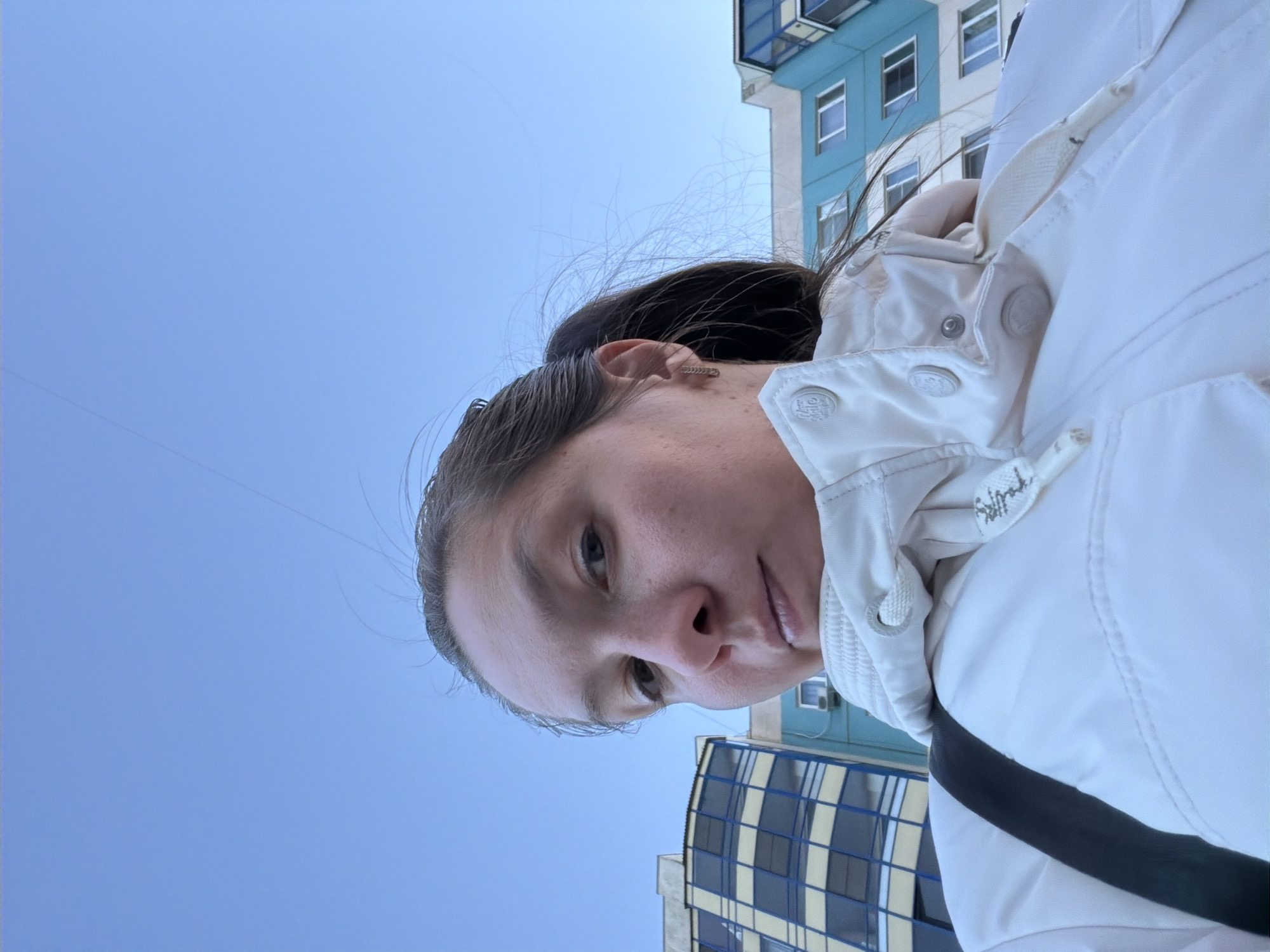 A woman stands outside in a city environment, likely in Unknown, Russia based on architectural details and the presence of Cyrillic script on the jacket drawstring. She wears a white puffer jacket and has dark hair pulled back. Buildings, one blue and the other with yellow and grey grid patterns, are visible in the background against a clear blue sky. The photo appears to have been taken at an upward angle, with the camera held below eye level. The woman is looking down slightly at the camera. A black strap, possibly from a bag, crosses her jacket.