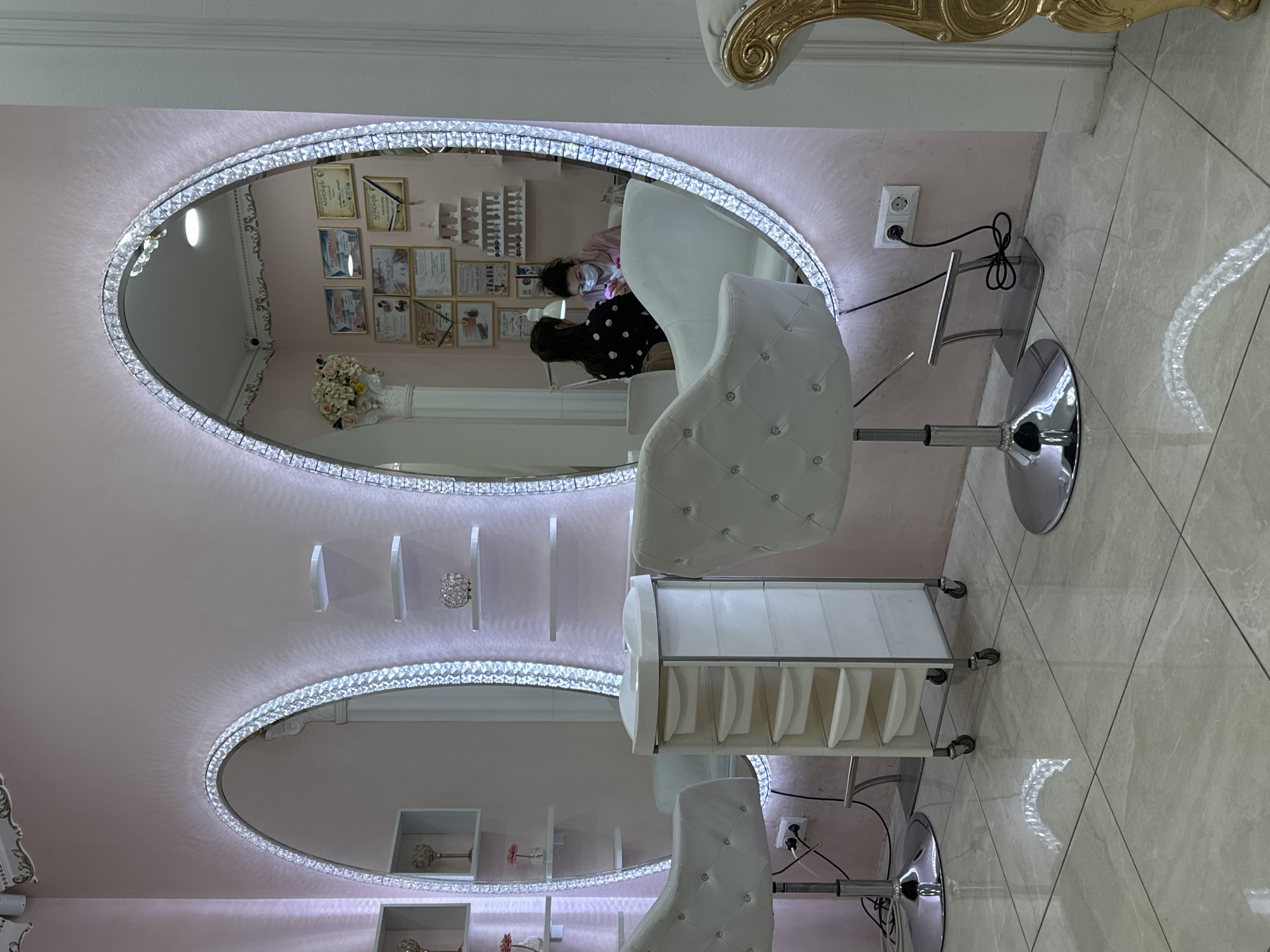 The image depicts the interior of a beauty salon, likely a nail salon or manicure studio. There are two large, oval-shaped mirrors with crystal or rhinestone embellishments surrounding the edges, which create a bright, diffused light. Reflected in the mirrors are various details of the salon, including framed certificates or licenses displayed on the wall, decorative shelves with small ornaments, and at least two individuals: one wearing a face mask and another with dark hair wearing a polka-dot shirt. The style of the salon is ornate and luxurious, indicated by the design of the white, tufted chairs. These chairs are adjustable salon chairs with chrome bases. Also in the frame is a white rolling cart, used to store supplies and equipment. The floor is tiled. The salon's decor is primarily pink and white. There is an electrical outlet and power cords visible on the wall. The location is presumed to be in Sofia, Bulgaria, although this is not visually confirmed in the image.
