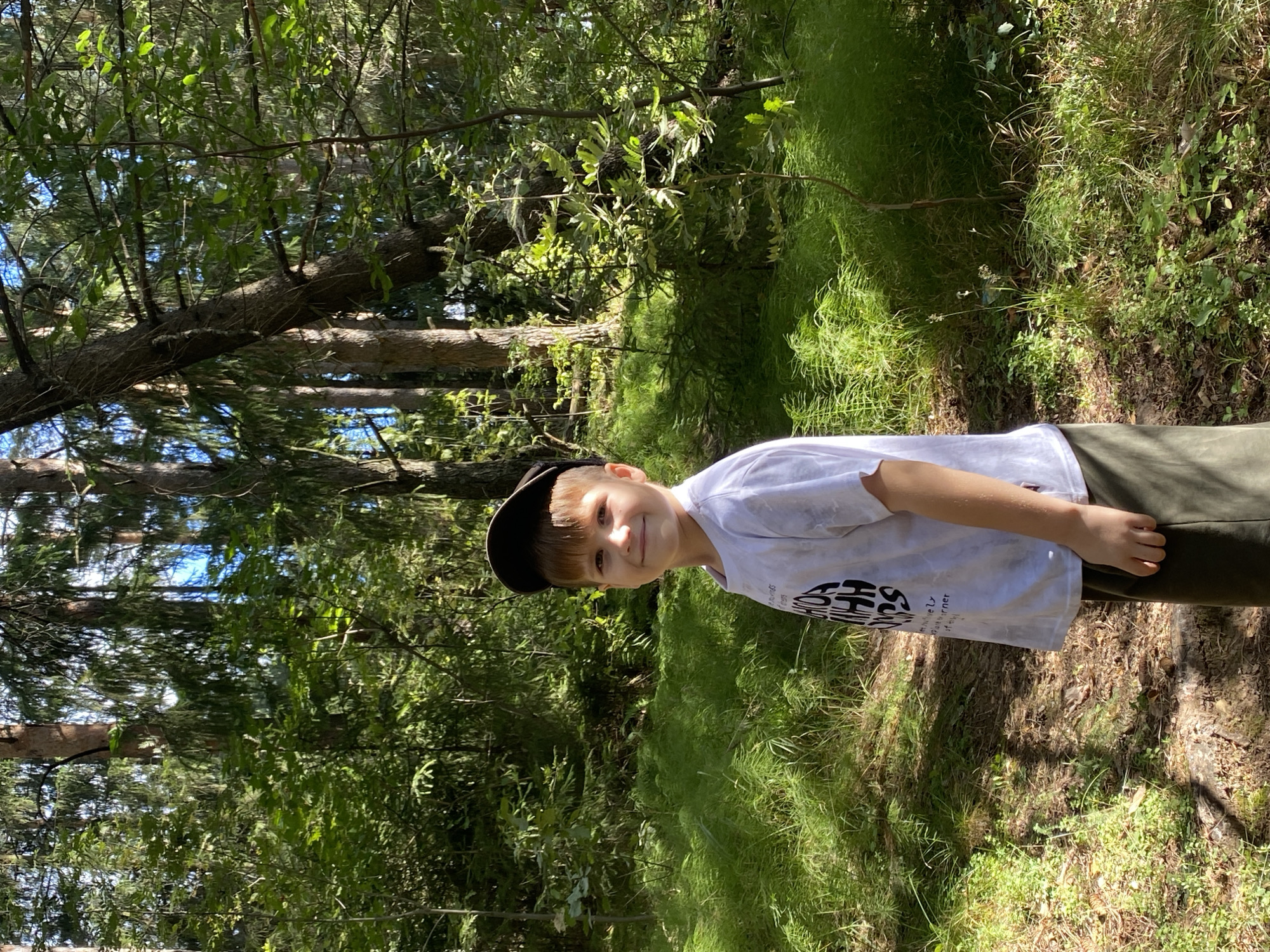 A young boy, wearing a white t-shirt and a dark baseball cap, stands in a forest. He is smiling and looking at the camera. The forest is lush and green, with tall trees and dense undergrowth. Sunlight filters through the leaves, creating a dappled effect on the ground. The boy is positioned slightly to the left of the frame, with the forest filling the rest of the image. The location is identified as Foki, Russia.
