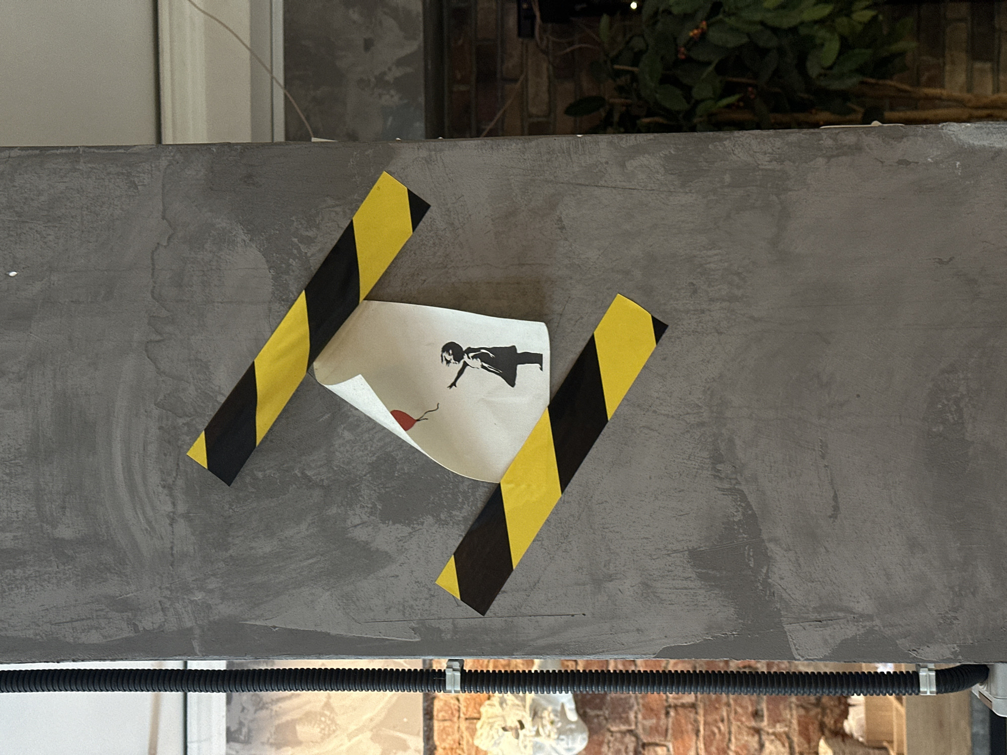 The image shows a section of a textured, gray concrete column in Moscow, Russia. Two strips of black and yellow hazard tape are affixed to the column. Between the tape strips, a piece of paper is attached, depicting a silhouette of a young girl reaching for a red balloon. The paper is slightly crumpled and appears to be a reproduction of Banksy's "Girl with Balloon" street art. A black, corrugated pipe runs vertically along the left side of the column, secured by a metal clamp. In the background, a brick wall and greenery are visible.