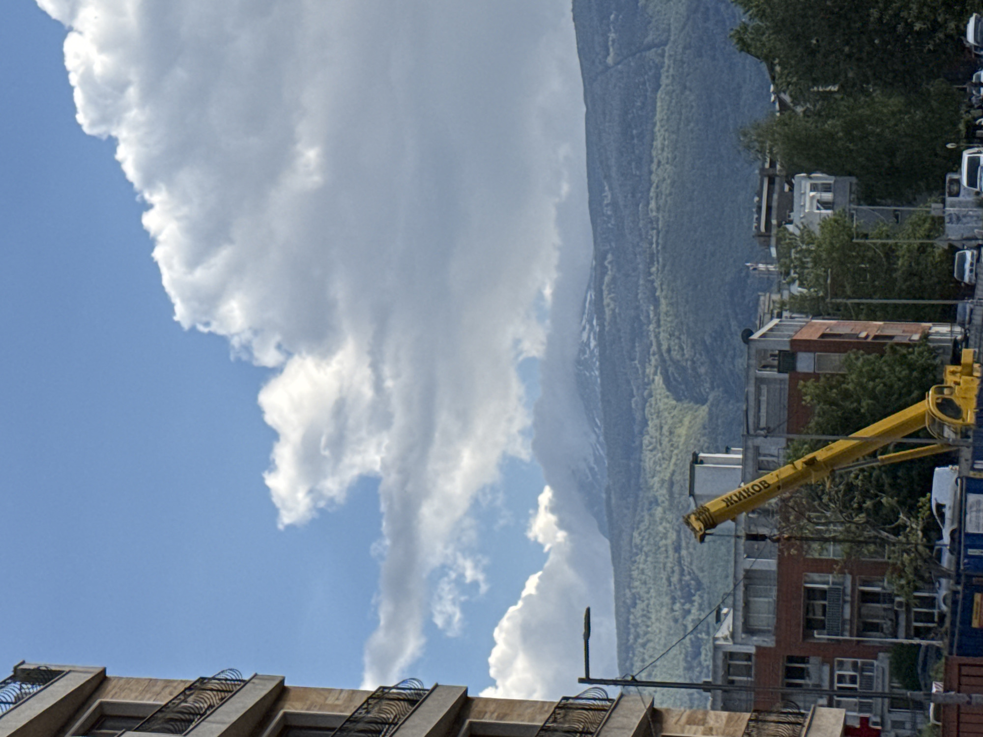 The image depicts a partly cloudy sky over a building in Sofia, Bulgaria, with a mountain range in the background. A yellow construction crane with the name "ZHIKOV" (ЖИKOВ) visible on its arm is positioned near the buildings. Trees are interspersed among the buildings and along the mountainside. Some vehicles are seen parked on a street near the buildings.