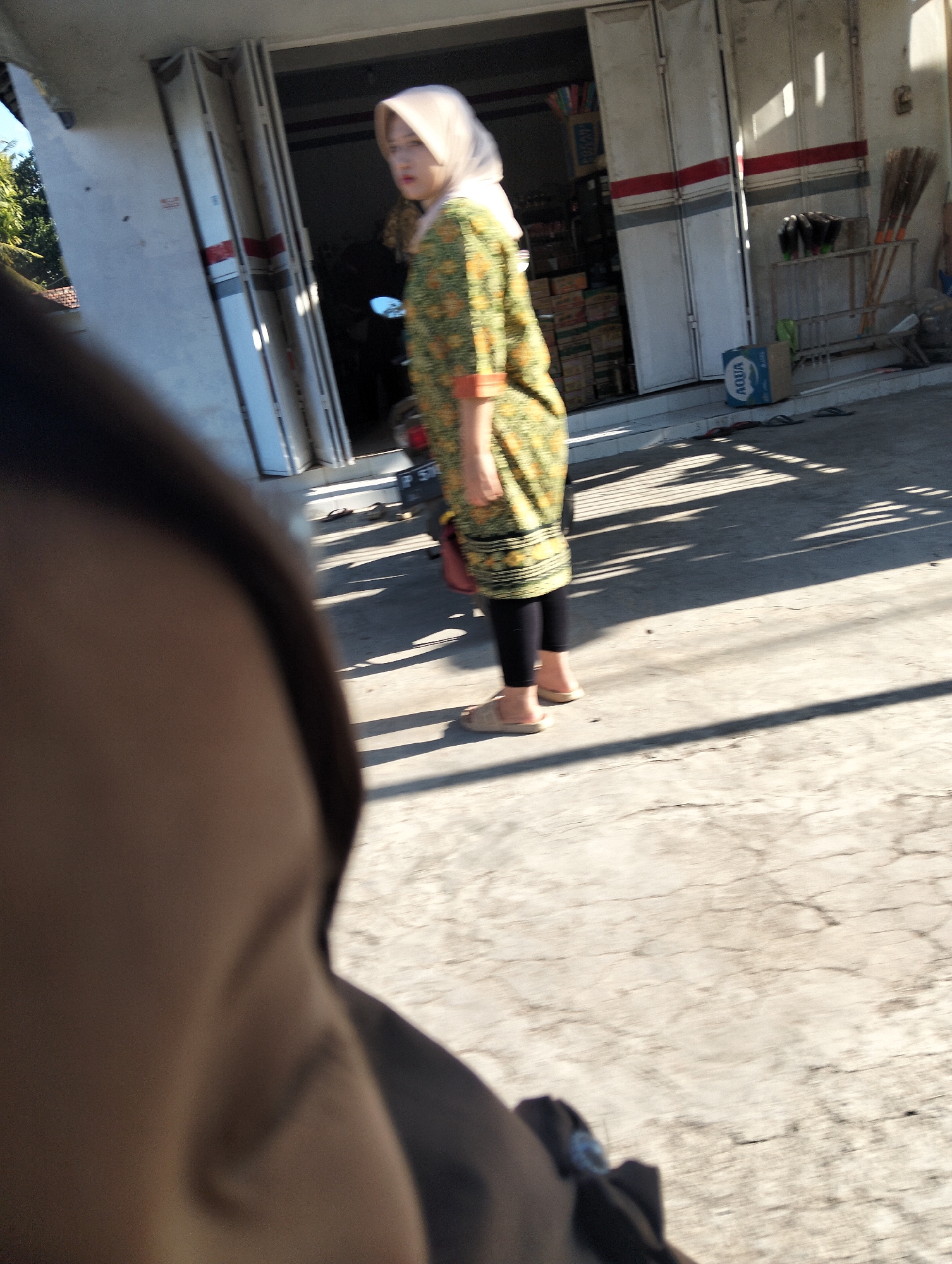 A person wearing a hijab and a colorful patterned dress walks on a concrete surface in front of a store with closed folding doors in Kedungringin, Indonesia. The person is wearing black leggings and light-colored slip-on sandals. Sunlight casts strong shadows on the ground. In the background, there are shelves with brooms and other items. A blue "Aqua" branded box is visible on the ground near the store entrance. A portion of a motorcycle is also visible. The foreground is blurred and appears to be a close-up of another person's face and shoulder.