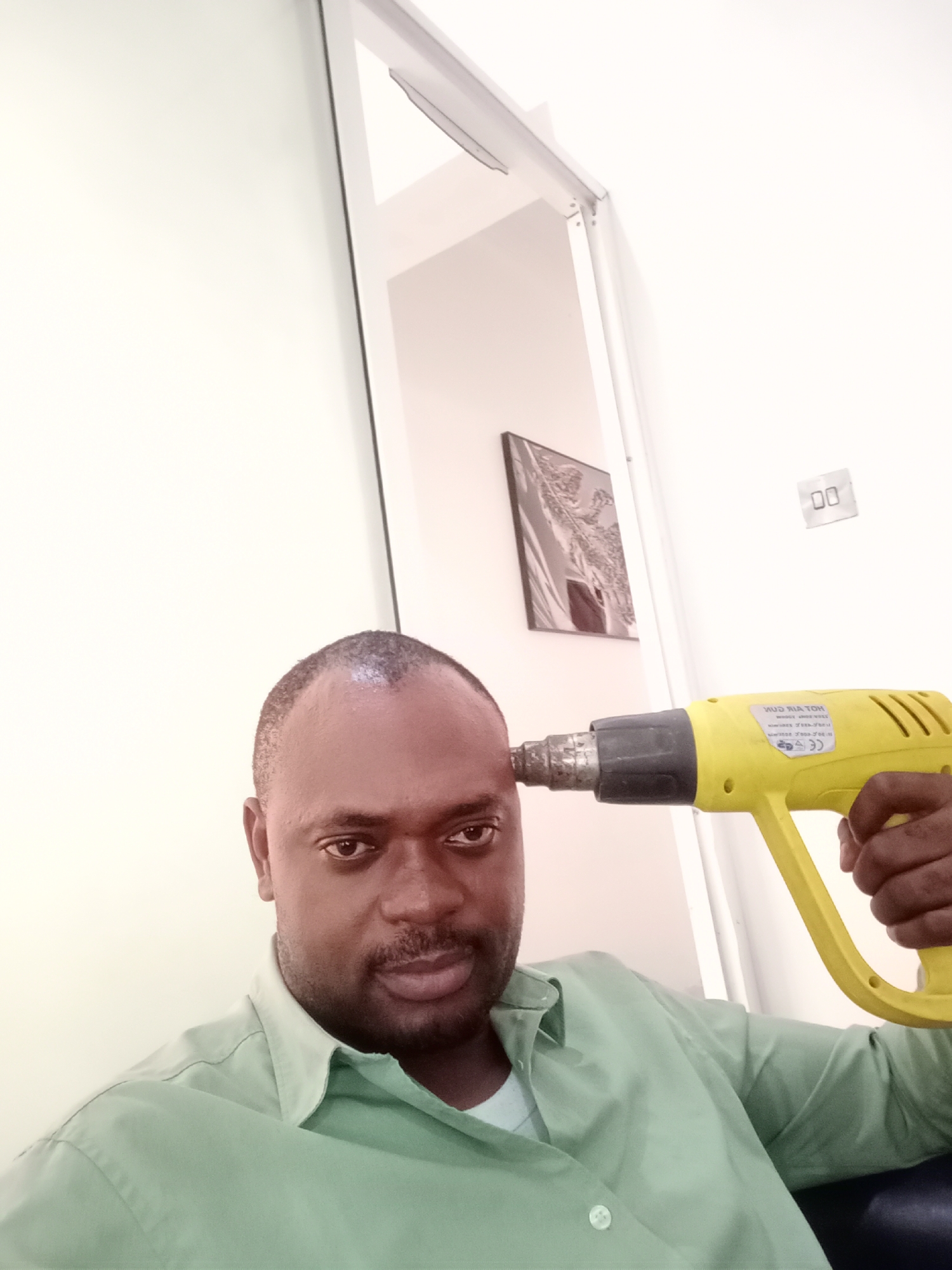 A man of African descent is depicted in a close-up, chest-up shot, holding a bright yellow heat gun to his forehead. He wears a light green collared shirt and has short dark hair and a beard, looking directly at the viewer with a neutral to slightly serious expression. His right hand grasps the handle of the heat gun, with the nozzle pressed against his left temple, creating an illusion of holding a pistol.
The background is a plain white wall, indicating an indoor setting. To the man's left, a light-colored doorframe is visible, suggesting an adjacent room. On the wall behind him, a framed picture, possibly an abstract design or a monochrome landscape, hangs vertically. Further right on the wall, an electrical outlet or switch cover with the numbers "00" printed on it is discernible.
Visible text on the yellow heat gun includes mirrored or partially obscured words that appear to be "HOT AIR GUN" and a "CE" mark, alongside other smaller, illegible text. The scene is well-lit, characteristic of an indoor environment, possibly an office or a modern home in Gwarinpa, Nigeria. The image captures a posed, possibly humorous or symbolic moment.