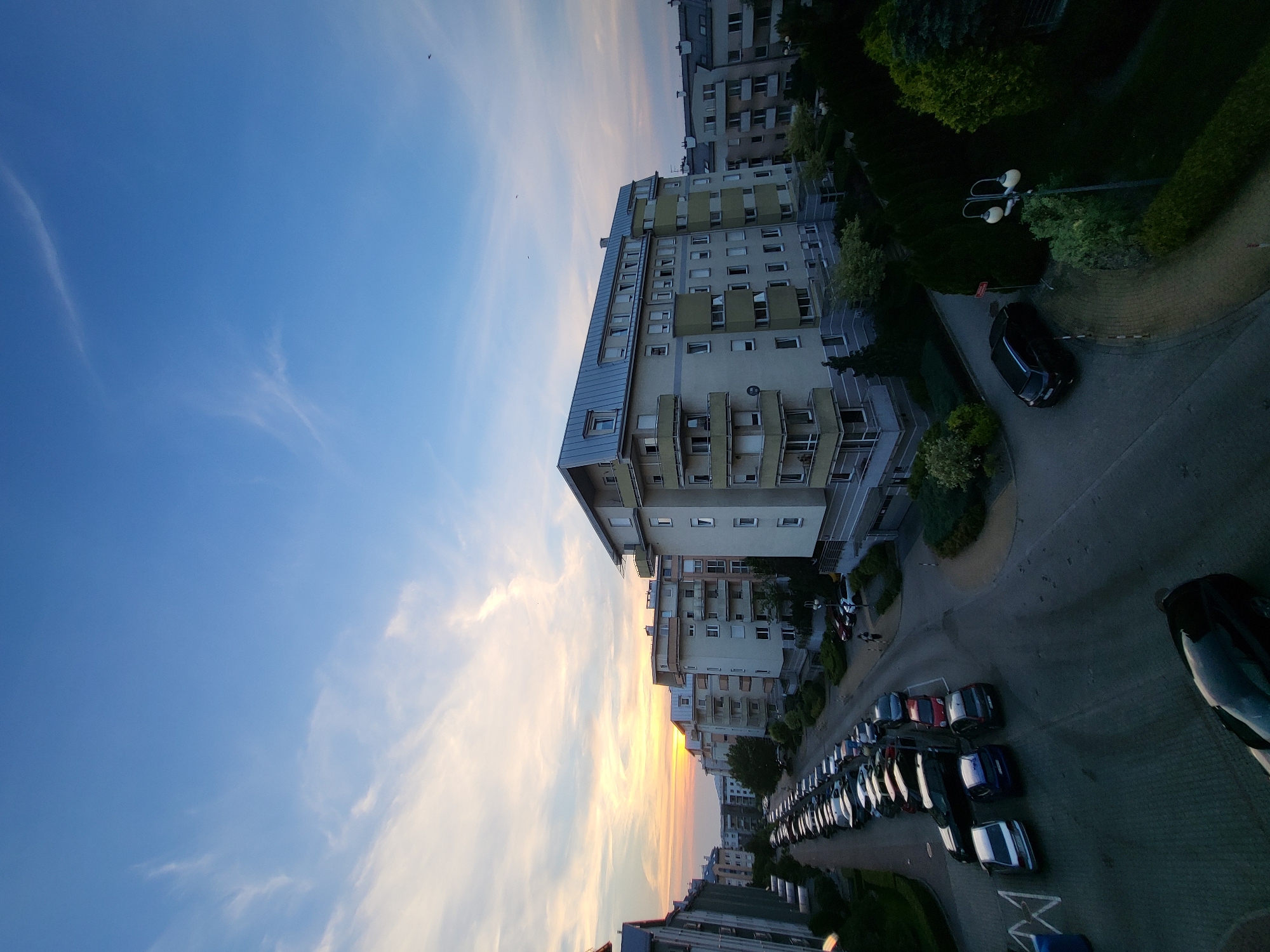 This image captures a view of apartment buildings and a parking lot at dusk. The sky is a blend of deep blue and soft orange and yellow hues, indicating either sunrise or sunset, with wispy clouds scattered across it. A few small birds are visible in the sky.
The main subjects are several multi-story apartment buildings with balconies and numerous windows. In the foreground and extending into the middle ground, a parking lot is filled with a variety of parked cars. Some of the cars are dark-colored, while others are lighter shades, including white, silver, and red. One black car is prominently parked near a street lamp.
The scene appears to be a residential area, possibly a housing complex. The time of day is clearly evening or early morning, given the twilight sky and the soft lighting. The weather seems calm, with no indication of rain or strong winds. The image's perspective is from a high vantage point, looking down onto the scene. The overall mood is serene and quiet, characteristic of the transition between day and night. There is no visible text in the image.
