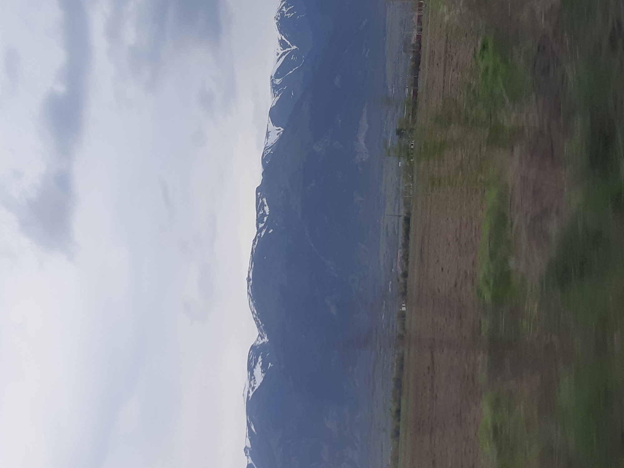 The image depicts a landscape with mountains partially covered in snow on the left, a field in the center, and greenery on the right. The sky is cloudy. Based on the presence of mountains and fields, this could potentially be Marino Pole, Bulgaria, however, there is no confirmation of buildings/infrastructure.