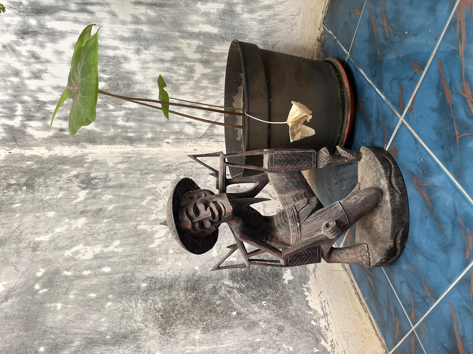 The image depicts an outdoor scene on a tiled floor. In the foreground, a dark wooden carving of a man wearing a conical hat and carrying a yoke on his shoulders is positioned to the left. The carving has a detailed, aged appearance with visible facial features and clothing. To the right of the carving is a dark plastic pot containing a green plant with large leaves and several thin stems. One dried, withered leaf hangs down from a stem inside the pot. The background consists of a rough, textured concrete wall with patches of white paint and grey discoloration, suggesting an aged or weathered surface. The floor is covered in blue tiles with reddish-brown veining. The scene appears to be an outdoor corner, possibly a patio or courtyard in Karangkebagusan, Indonesia.