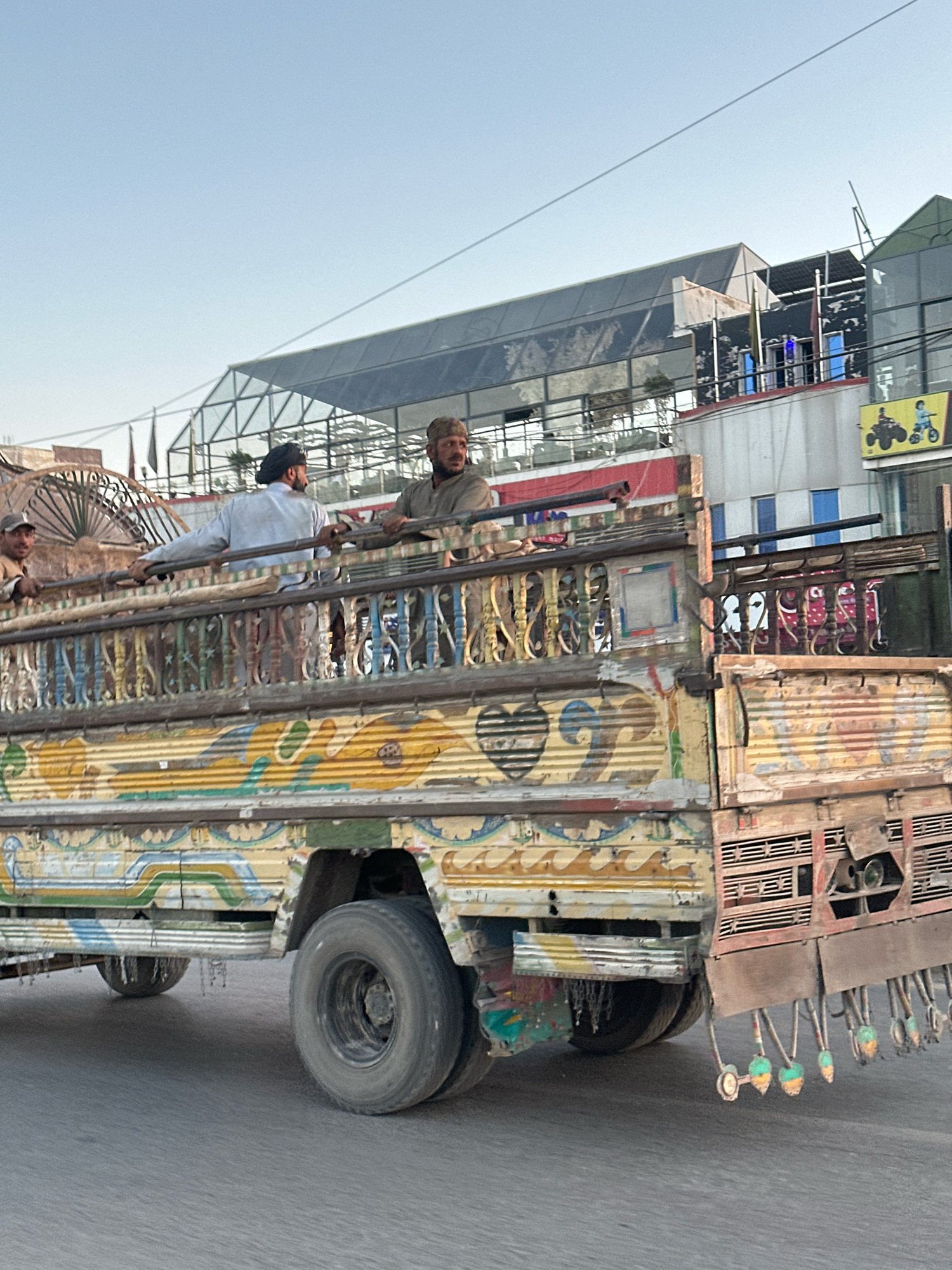 A slightly low-angle, medium shot captures a vibrantly decorated, weathered truck with several men standing on its bed, traveling on a paved road under a clear, pale blue sky. The truck, predominantly red, yellow, and blue, features ornate, colorful paintwork on its sides, including floral motifs and what appear to be heart shapes. The truck bed is lined with a metal railing, also painted, and has a metal grate at the rear from which several decorative, bell-shaped objects hang.
Three men are visible. Two are standing on the truck bed, leaning on the railing and looking to the right. One man wears a light-colored shirt and a dark turban. The other wears a light brown, button-up shirt and a cap. A third man, wearing a lighter cap and shirt, is partially visible on the left side of the truck, also leaning on the railing.
In the background, beyond the truck, is a multi-story building with a modern, angular glass and metal facade, reflecting the sky and surrounding structures. Parts of the building appear to be under construction or undergoing renovation, with exposed metal beams and unfinished sections. A sign featuring cartoonish images of people on ATVs and motorcycles is visible on the right side of the building. The overall environment suggests an urban setting in Quetta City Tehsil, Pakistan, likely during daylight hours, given the clear sky and shadows.