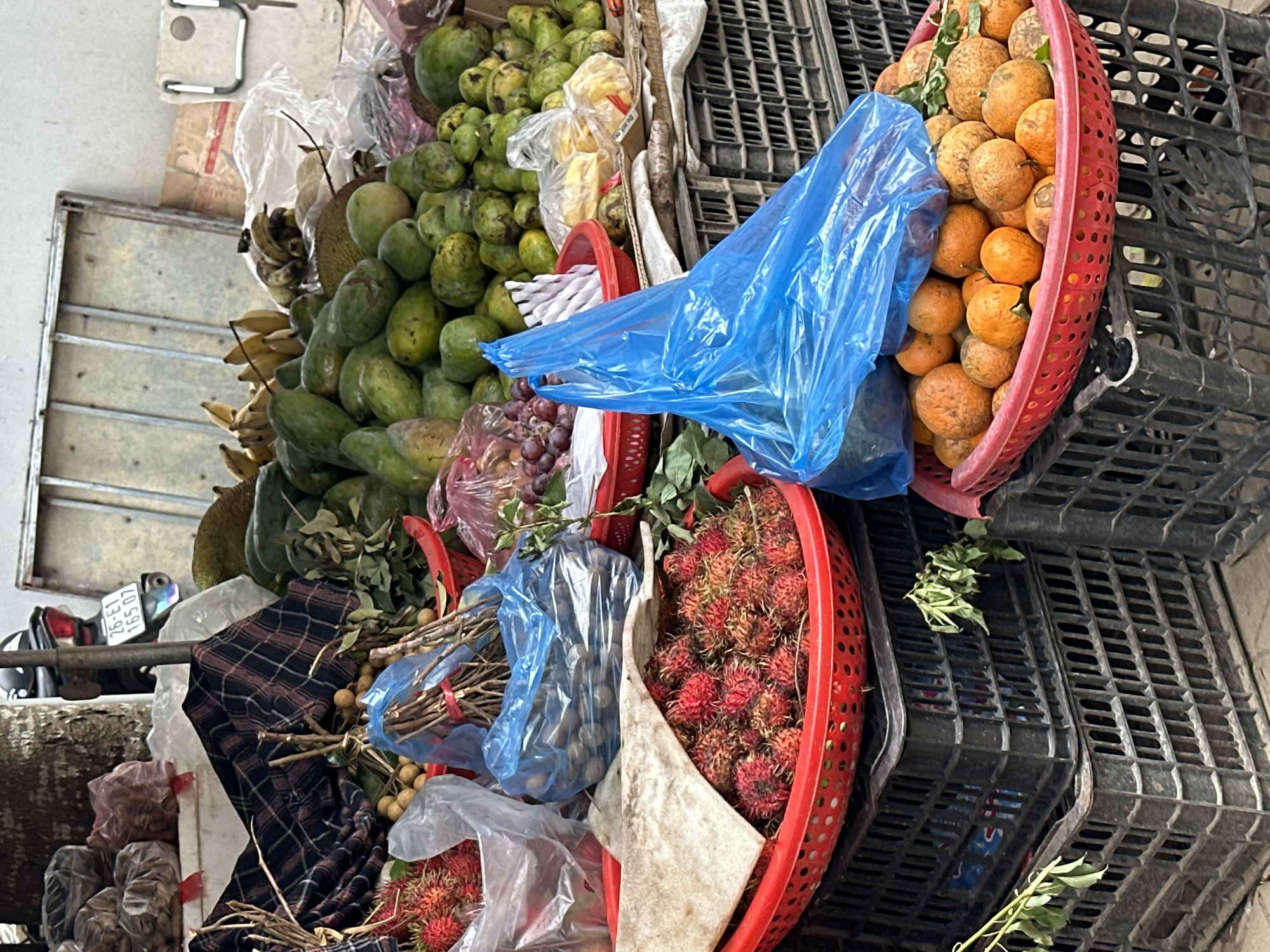 A close-up, eye-level shot captures a fruit stand at a street market in Bắc Yên, Vietnam. The focus is on a colorful display of various fruits, predominantly in plastic bags and red baskets. In the foreground, a red basket overflows with round, orange fruits, likely tangerines or small oranges. To the left, another red basket holds a cluster of red, spiky fruits, identifiable as rambutans, with a piece of light-colored cloth draped over them. Behind these, a blue plastic bag, partially obscuring the view, sits atop a stack of black plastic crates. Further back, a larger variety of green fruits, possibly mangoes and papayas, are piled high. Bunches of yellow bananas and some dark red grapes in a red bowl are also visible, interspersed with more plastic bags and bundles of dry sticks. The background reveals hints of the market environment, including a motorcycle with a license plate, metallic shelving, and a textured wall. The overall impression is a vibrant, bustling scene of commerce.