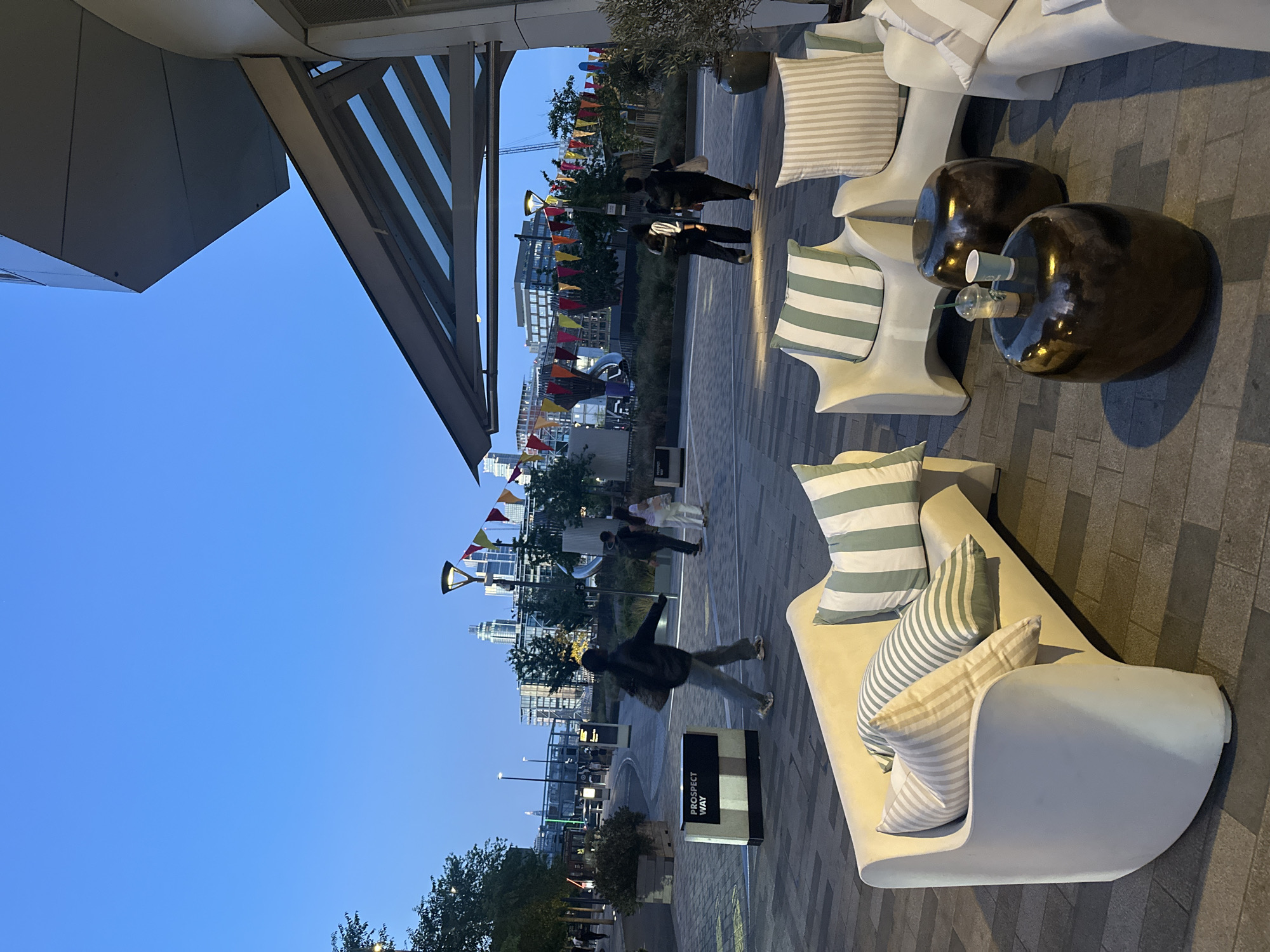 The image depicts an outdoor area in London, United Kingdom, featuring modern, white lounge furniture with striped pillows arranged on a paved surface. In the background, a cityscape with a suspension bridge and bunting strung overhead suggests an urban environment. People are visible walking in the distance, while the foreground is dominated by the seating arrangement and its immediate surroundings. A sign reading "PROSPECT WAY" is also visible, indicating a street name. The sky is a gradient of blue, suggesting either dusk or dawn.