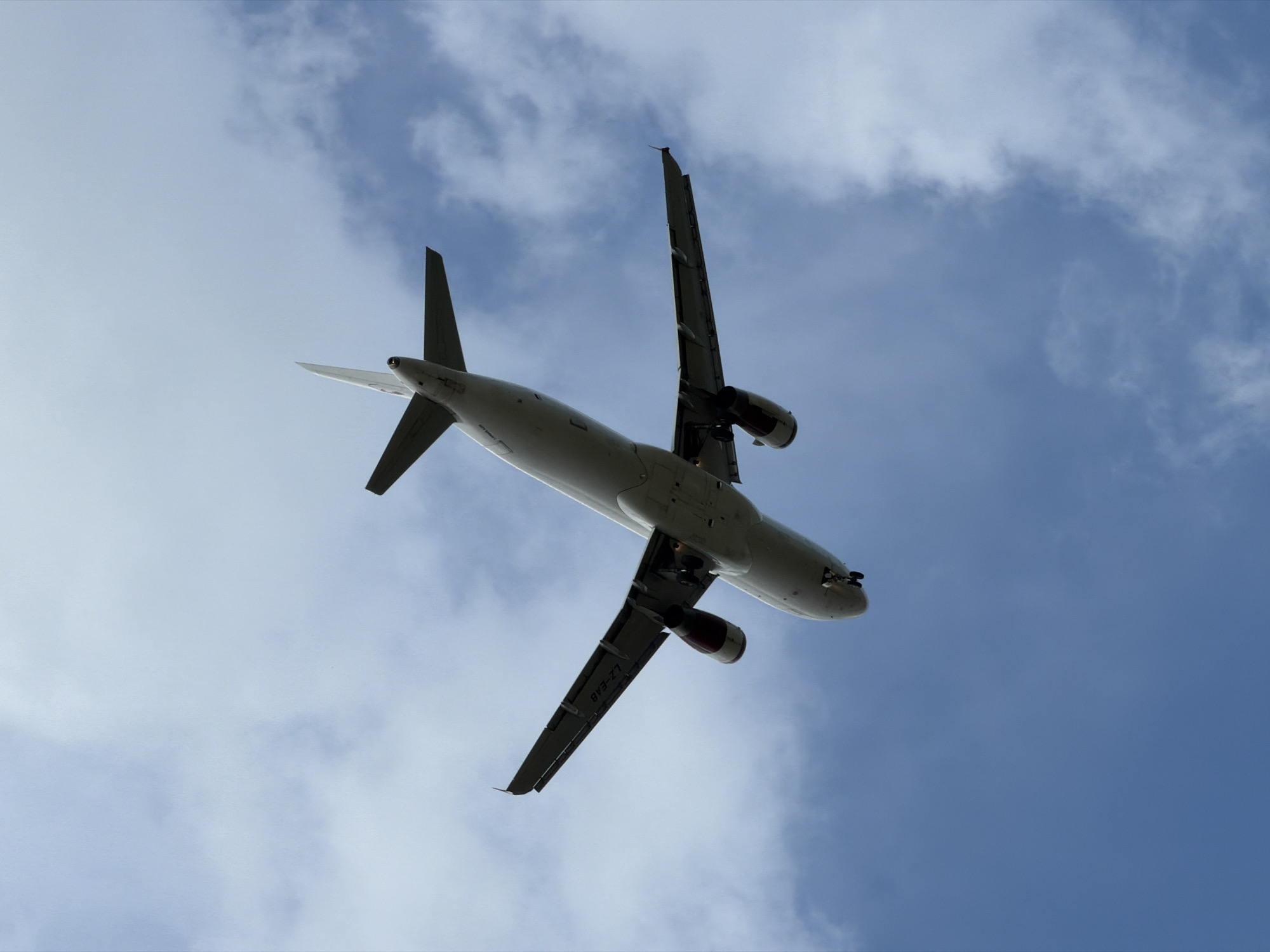 An airplane is captured from below, flying through a cloudy sky over Burgas, Bulgaria. The underside of the aircraft is visible, including its wings, engines, and landing gear. The airplane's registration number, LZ-EAB, is faintly discernible on one of the wings. The sky is a mixture of blue and white clouds, suggesting it is daytime. The perspective from below emphasizes the vastness of the sky and the imposing presence of the aircraft.