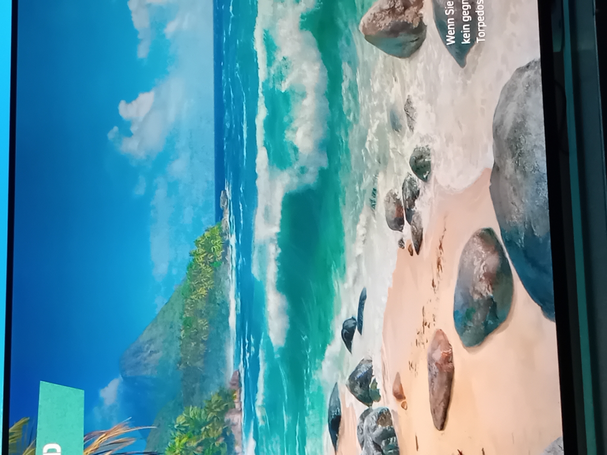 The image displays a vibrant, picturesque tropical beach scene, likely shown on a digital screen, as indicated by visible black borders on the right and top, and a light blue border on the left. The scene is dominated by a clear, turquoise ocean with prominent white waves crashing onto a light beige sandy beach. Numerous smooth, dark and light-colored rocks and pebbles of various sizes are scattered across the wet and dry sand.
On the left side of the frame, a lush green island or coastal landmass is visible, covered in dense vegetation, including palm-like trees, and featuring a steep, mountainous peak in the background. The sky above is bright blue with scattered white, fluffy clouds, suggesting a sunny day, likely late morning or early afternoon.
No human subjects or specific activities are visible; the image captures a serene, natural landscape. The movement of the waves is the primary activity depicted. A partial green rectangular element is visible in the bottom-left corner of the screen, and a dark element is in the bottom-right.
Visible white text is overlaid on the sandy beach area on the right side of the image. The partial text is in German and reads: "Wenn Sie" (If you), "kein gegn" (likely part of "kein Gegner" - no opponent), and "Torpedos". This suggests the display might be from a game, an advertisement, or informational content.
While the location context provided is Figueira da Foz, Portugal, the visual elements such as the vibrant turquoise water, specific tropical-looking vegetation, and mountainous island formation do not typically align with the geography of Figueira da Foz, which is situated on the Atlantic coast. The scene itself strongly evokes a generic tropical paradise rather than a specific European coastal landscape.