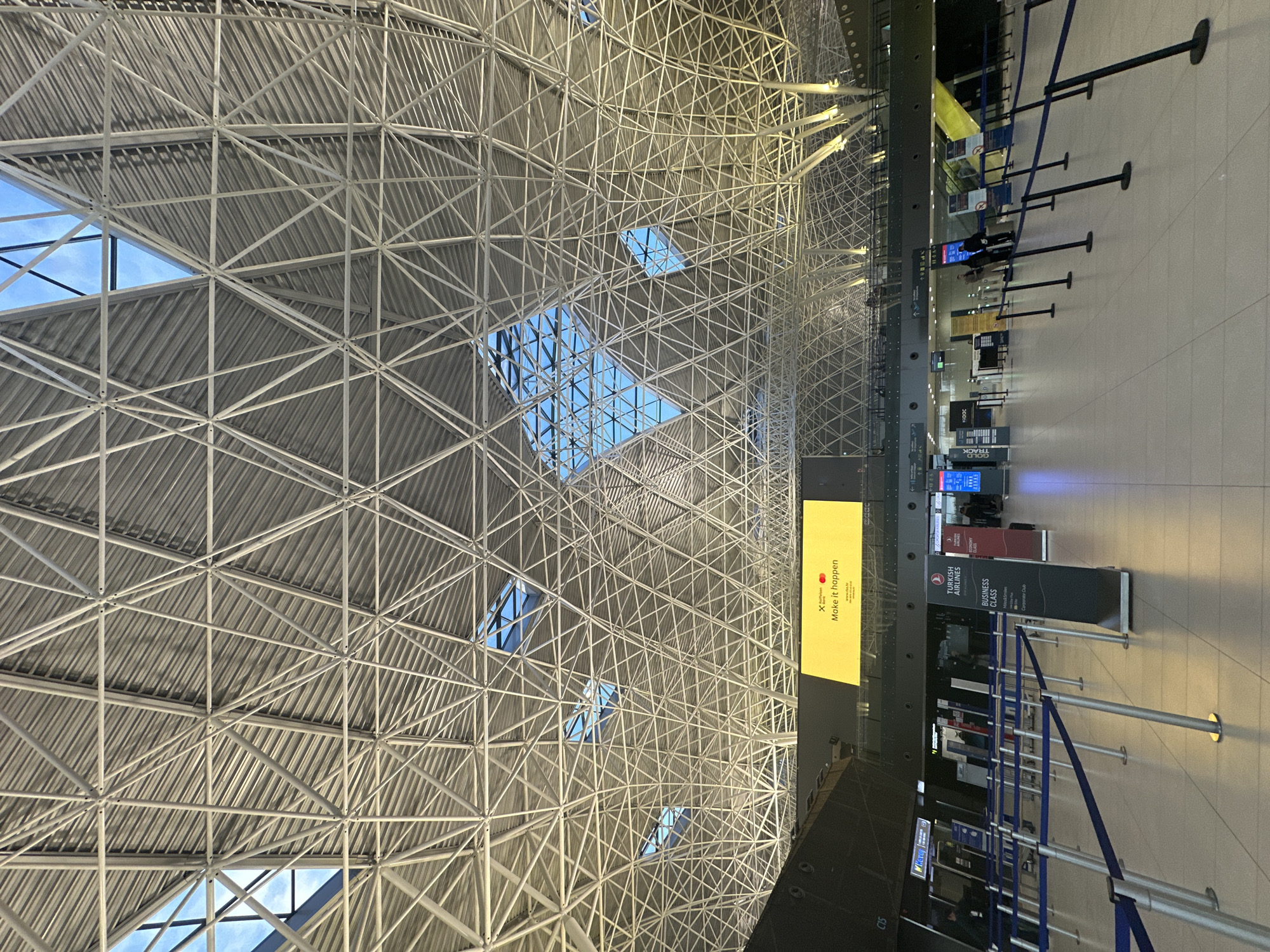 The image shows the interior of Zagreb Airport (ZAG), located in the city of Velika Gorica, Croatia. The view is looking upwards, showcasing the complex structural roof with a network of white steel beams and triangular skylights allowing natural light to enter the building.
Below the roof, on the right side of the image, is a section of the airport terminal. It displays a series of check-in counters or boarding areas. There are signs indicating "Turkish Airlines Business Class" and an advertisement for "Make it happen". Queue barriers, marked by blue straps and metal posts, are visible, guiding passenger flow. The floor is a light-colored tile or concrete. Several digital displays with flight information are present above the counters.