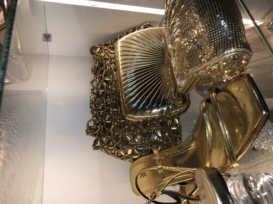 The image displays a close-up view of five golden fashion accessories arranged on a clear glass shelf within what appears to be a display cabinet. The specific location in Tema, Ghana, is not discernible from the visual data provided.
Subjects and Objects:
- One pair of golden high-heeled sandals is positioned on the left, partially obscured. The brand "ZARA" is visible on the insole of the right sandal. The sandals feature thin straps and a square toe.
- Three golden clutch bags are visible:
- A large, textured clutch with an intricate open-weave or beaded pattern is situated at the rear-center.
- A shiny, metallic, shell-shaped clutch with prominent radial grooves is centrally located in front of the textured clutch.
- A rectangular clutch with a fine metallic mesh or sequined surface, featuring a line of clear, faceted stones or rhinestones along its top edge, rests on the right side.
- Small, clear, irregular-shaped objects, possibly decorative acrylic pieces or bubble wrap, are present beneath the sandals and other items, likely for display or cushioning.
Environment:
The items are arranged on a clear glass shelf. Another clear glass surface, possibly an upper shelf or the top of the display case, is visible at the top of the frame, reflecting light. The background consists of white or light-colored paneling, suggesting an enclosed display area. Artificial lighting illuminates the scene, creating reflections and highlights on the metallic surfaces of the accessories. No people are visible, and no actions or interactions are depicted.