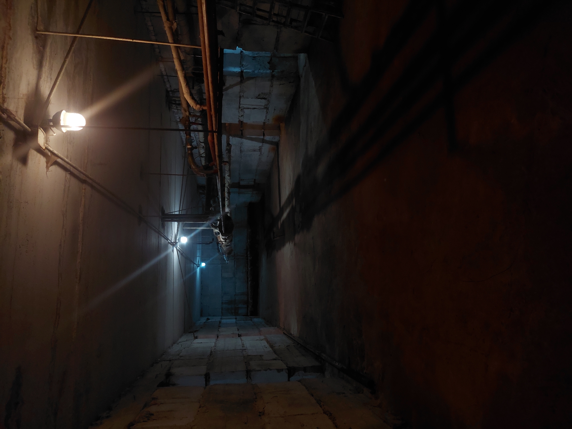 The image depicts a dimly lit, subterranean passage. The walls are concrete and show signs of wear and aging. A series of pipes and conduits run along the ceiling. The floor is paved with rectangular blocks, also seemingly made of concrete or stone. Several light fixtures with yellow bulbs are mounted on the walls, providing sporadic illumination. The perspective suggests a long, narrow corridor, and the lighting is such that the far end of the passage is shrouded in darkness. The image does not include any people. Without further information, it is not possible to confirm the location in Nadym, Russia.