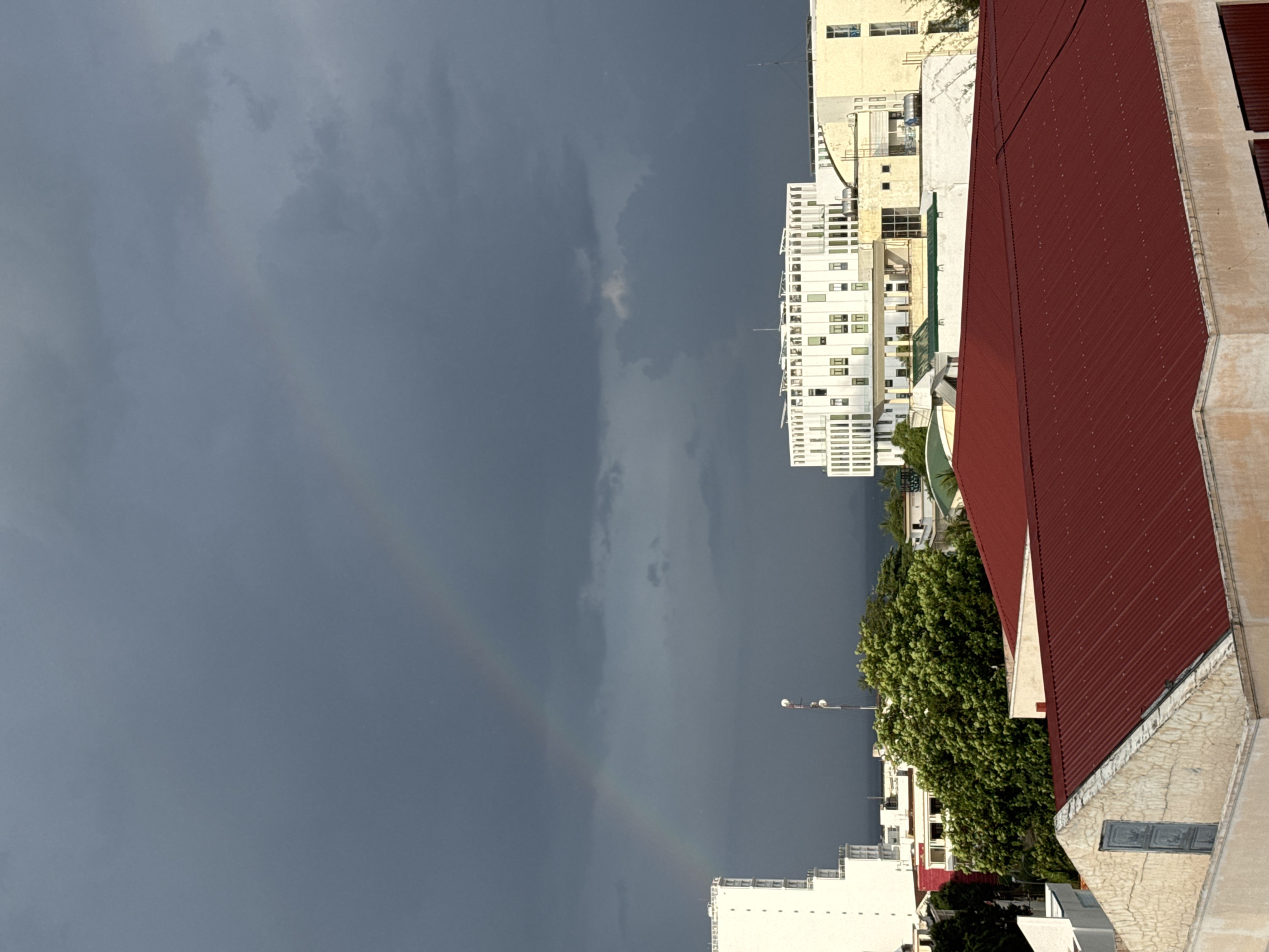 This image depicts a cloudy sky with a faint rainbow arching across it over the cityscape of Nha Trang, Vietnam. In the foreground, two buildings with red, corrugated metal roofs are visible. Behind them, a dense green tree is situated. Further back, a tall, white, multi-story building with many windows stands to the left. To the right of that, another building complex with a white, modernist facade and several floors with numerous windows is prominent. In the background, the ocean can be seen beyond the buildings, under the dark, overcast sky, suggesting recent or impending rain. The overall scene captures a moment of natural beauty against an urban backdrop in Nha Trang.