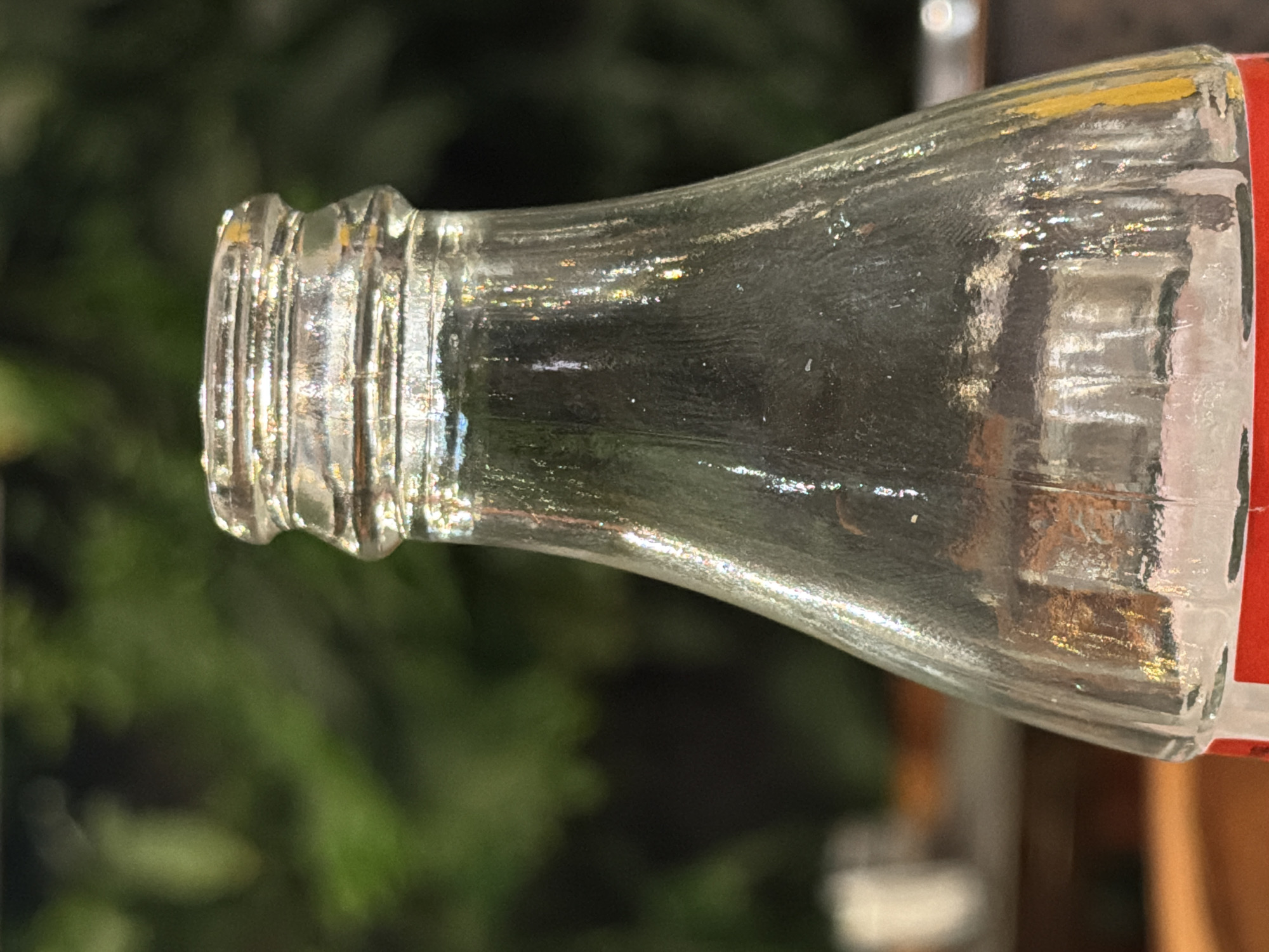 A close-up, horizontally oriented shot captures the neck and upper body of a clear glass bottle against a blurred green background. The bottle, possibly a Coca-Cola bottle due to the visible fragment of a red label on its right side, is made of textured, transparent glass. Bright highlights and iridescent reflections shimmer across its surface, particularly on the threaded neck and the curved body, indicating strong daylight.
The background is dominated by soft-focus green foliage, suggesting an outdoor setting such as a garden, park, or a view through a window. Further in the background, out-of-focus lighter elements and darker shapes hint at other structures or objects, possibly part of a building or furniture. No specific activity is depicted, and the bottle appears stationary. There is no discernible readable text on the partial label. While the location context of Sofia, Bulgaria is provided, the image itself does not contain any visual cues or landmarks to specifically confirm this geographical detail. The lighting implies bright, clear weather conditions.